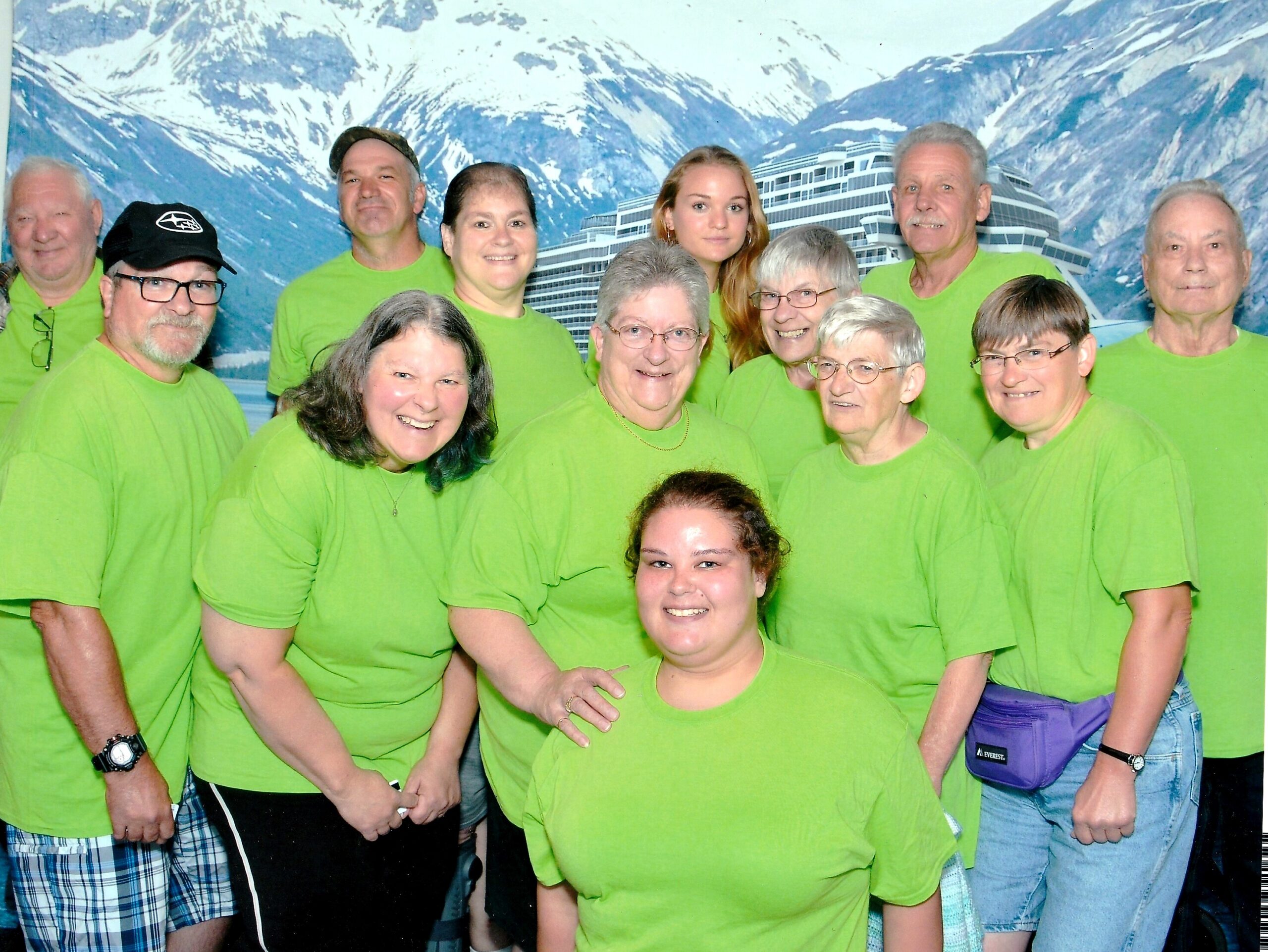 Group of twelve adults wearing bright green T-shirts posing for a group photo in front of a snowy mountain backdrop resembling a cruise ship scene.
