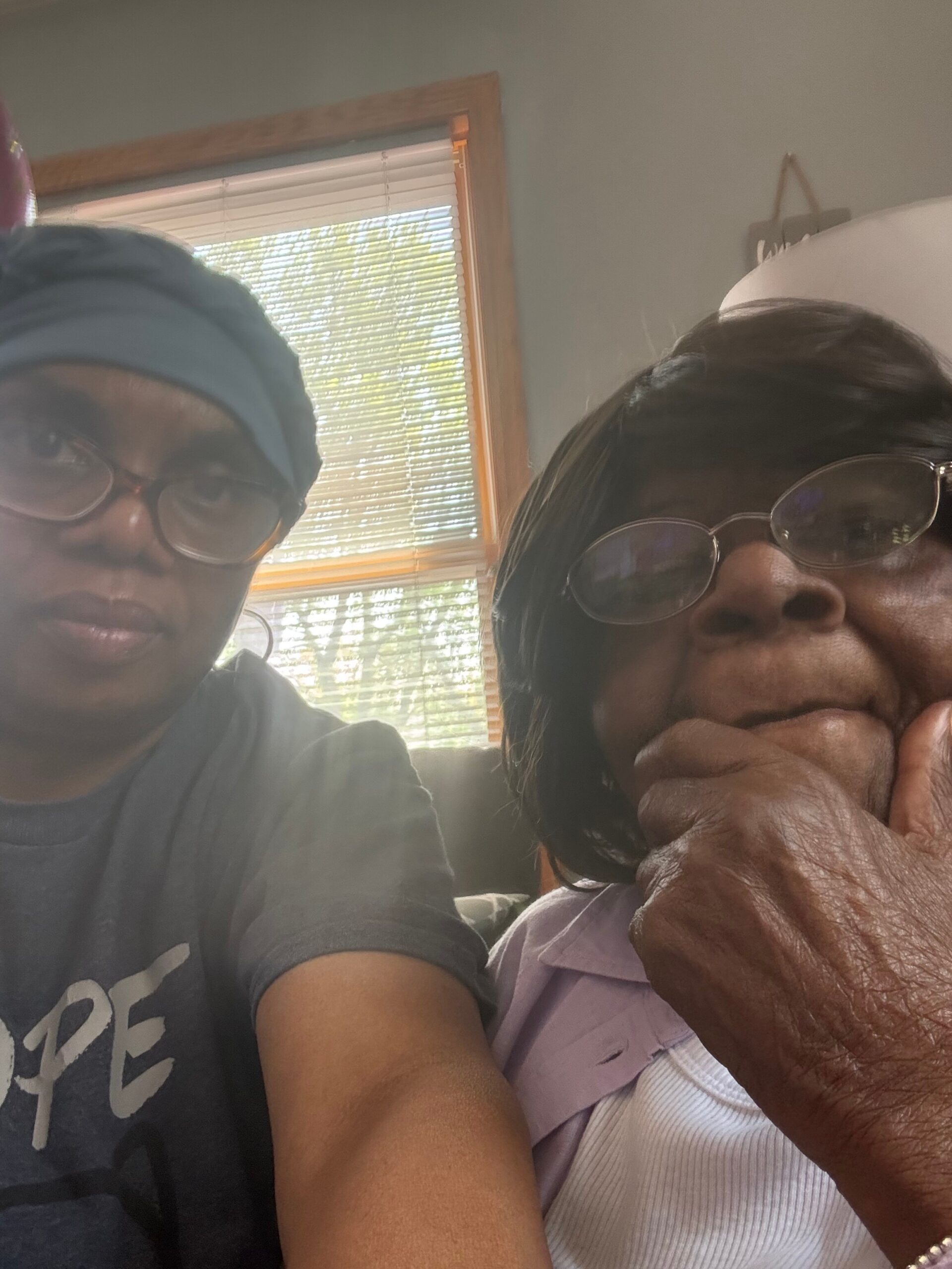 Two people posing for a close-up selfie indoors, one wearing a blue headband and glasses on the left, the other an older person with glasses on the right, hand near mouth; window blinds in the background.