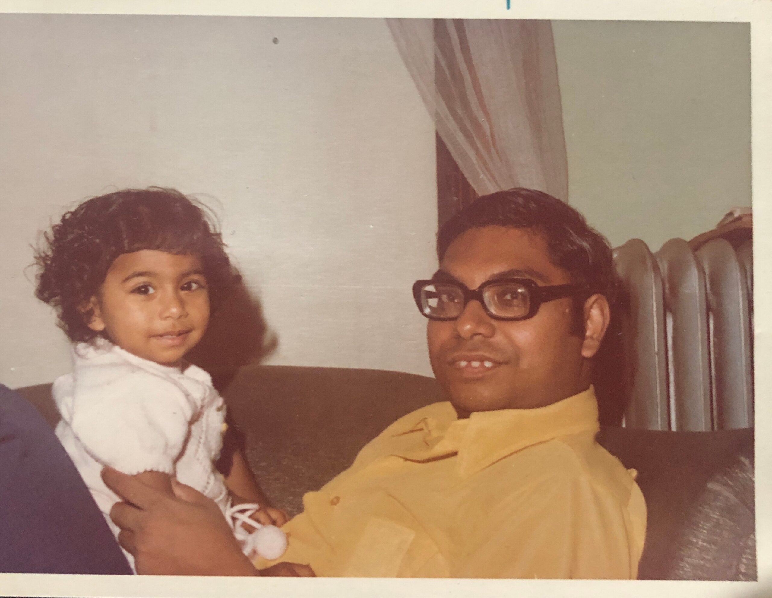 Man with glasses in a yellow shirt sits on a couch beside a young girl in a white dress, both looking at the camera in a living room.