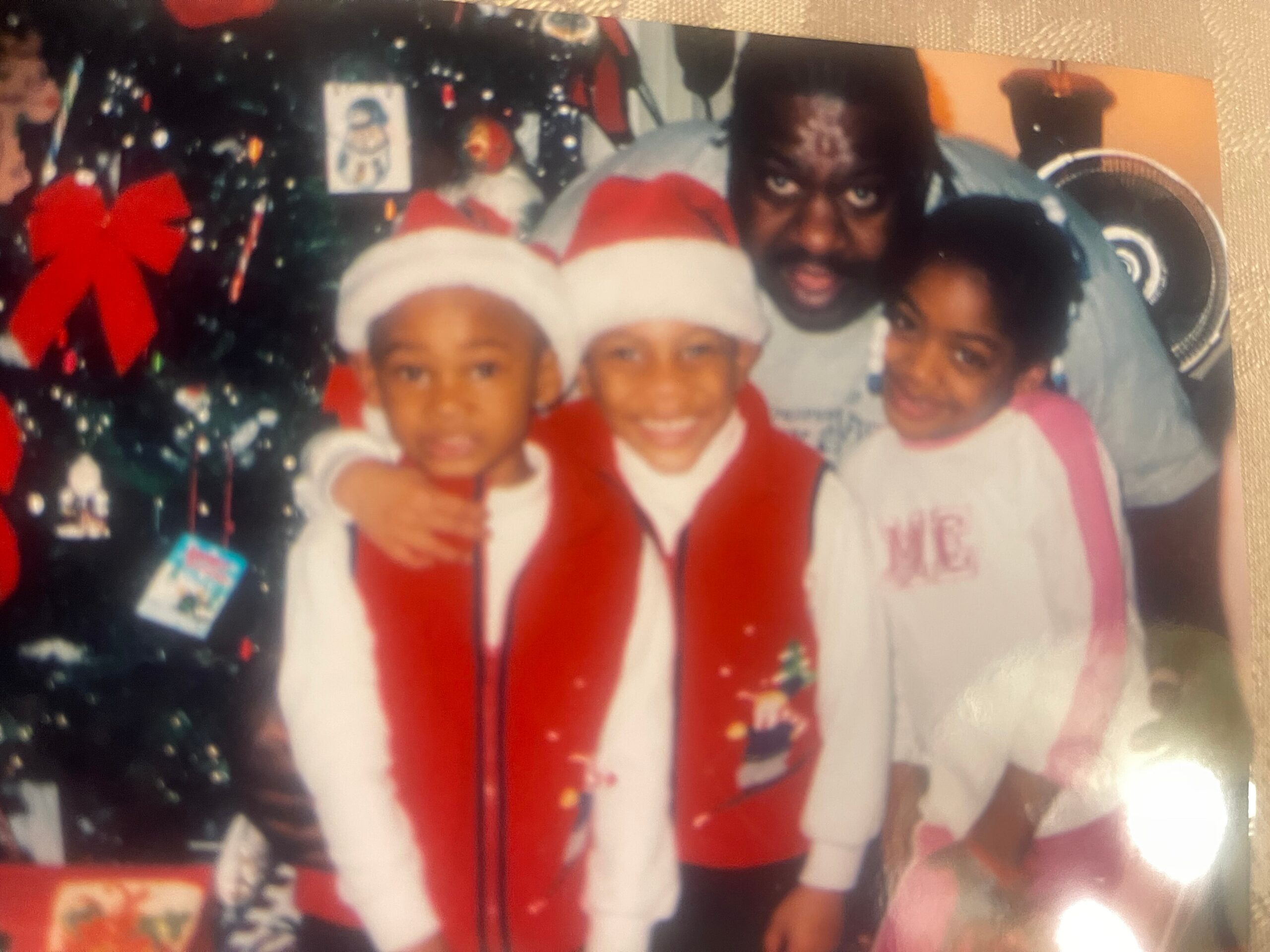 Family photo: four people in holiday attire posing together, wearing Santa hats, with a decorated Christmas tree in the background.