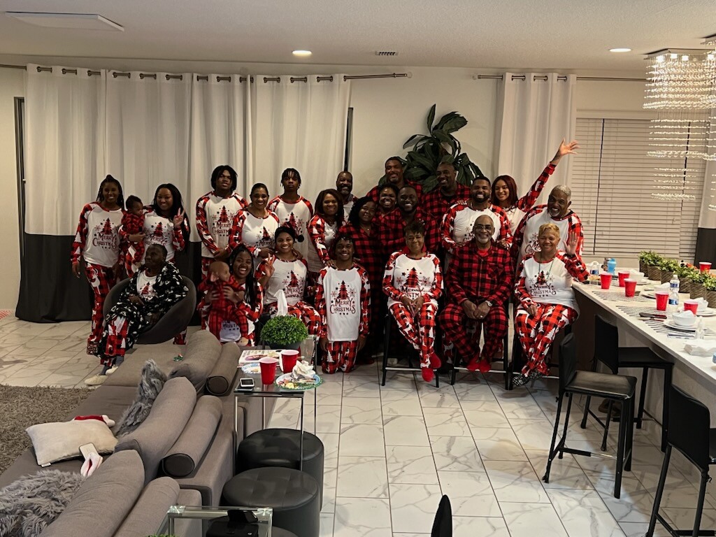 Group of people in matching Christmas-themed pajamas pose for a group photo in a bright living room with a Christmas party setup at a long table and couch area behind them.