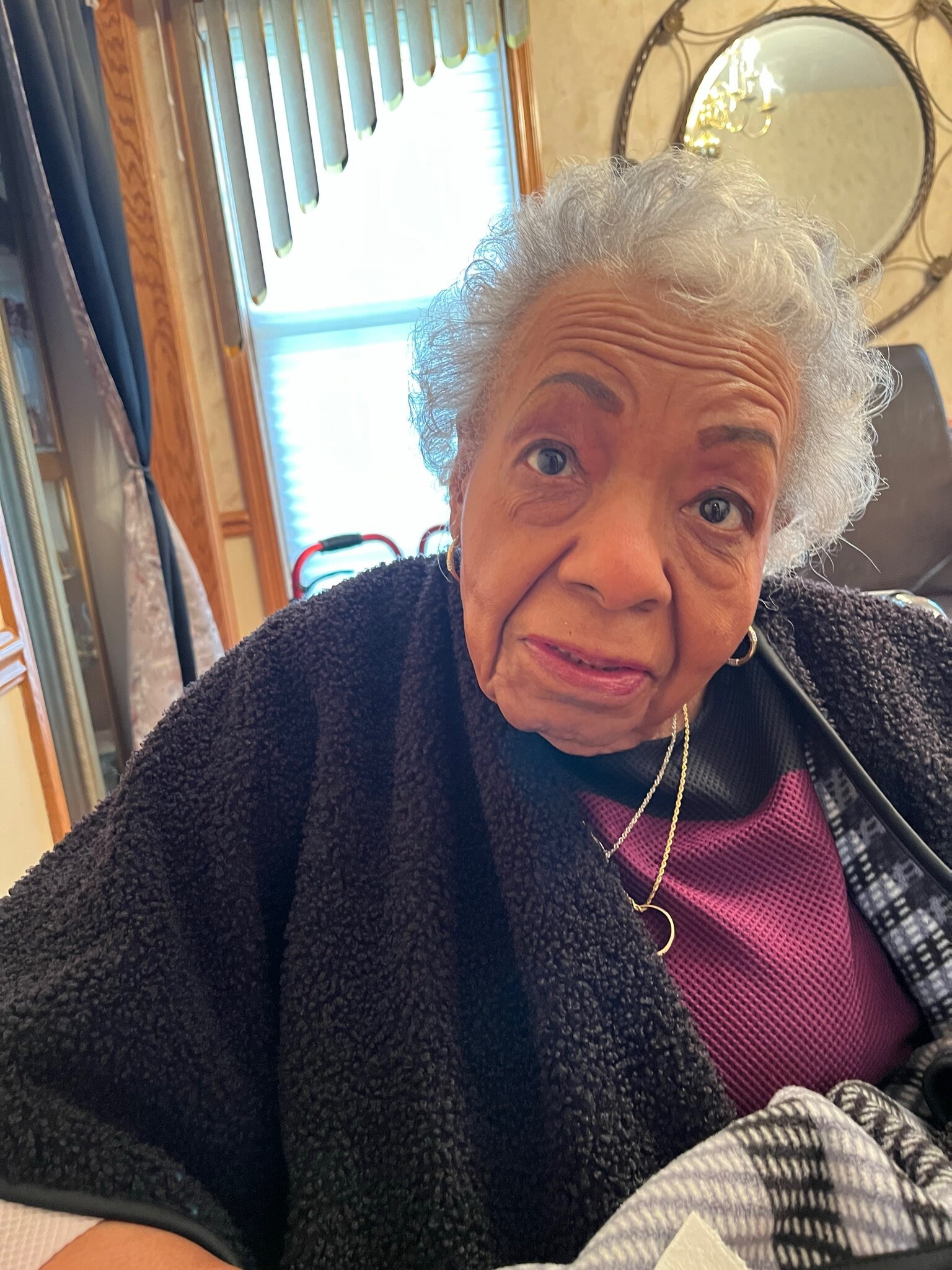 Close-up selfie of an elderly woman with gray hair, wearing a pink top and black sweater, in a cozy room with wood trim and a round mirror.