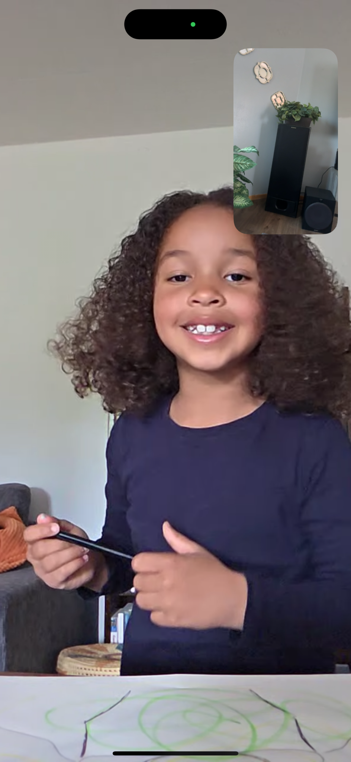 Happy young girl with curly hair drawing with a pencil at a table; green shapes on white paper, wearing a dark shirt; inset shows a plant and speaker in another room.