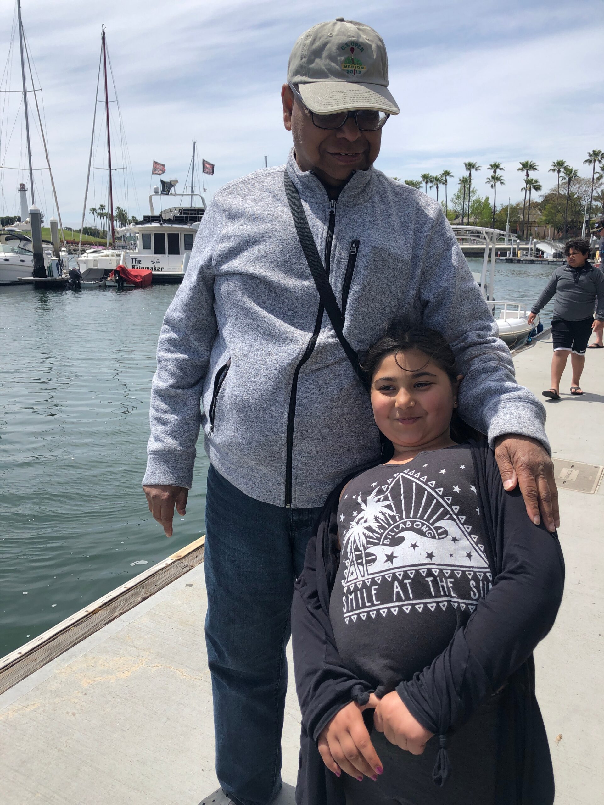 Man in a gray zip-up jacket and cap stands on a marina dock with a girl in front of him, boats and palm trees in the background.