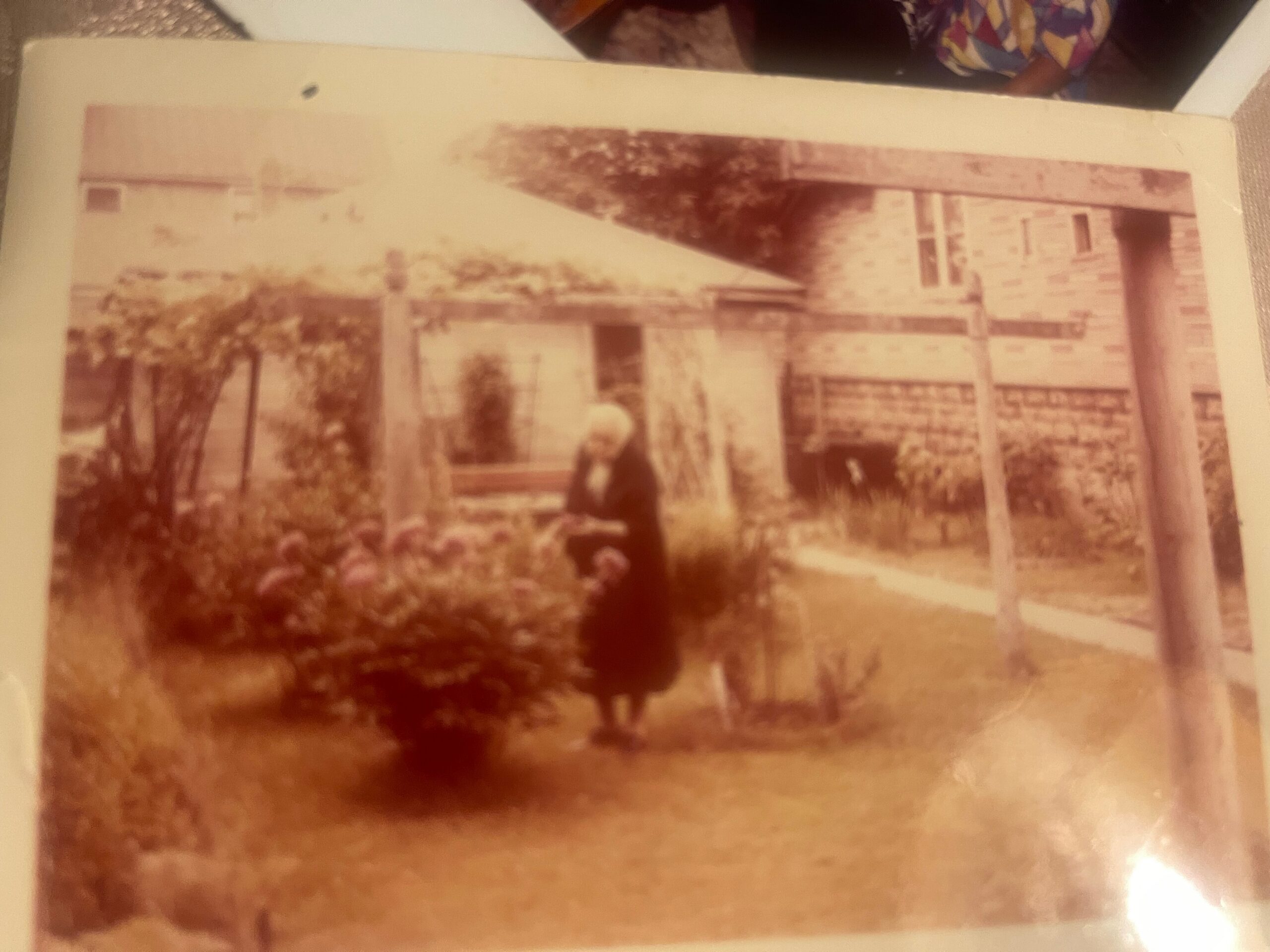 Old sepia-toned photograph of a brick house with a garden in front; a person stands near the foreground, partially blurred by age.
