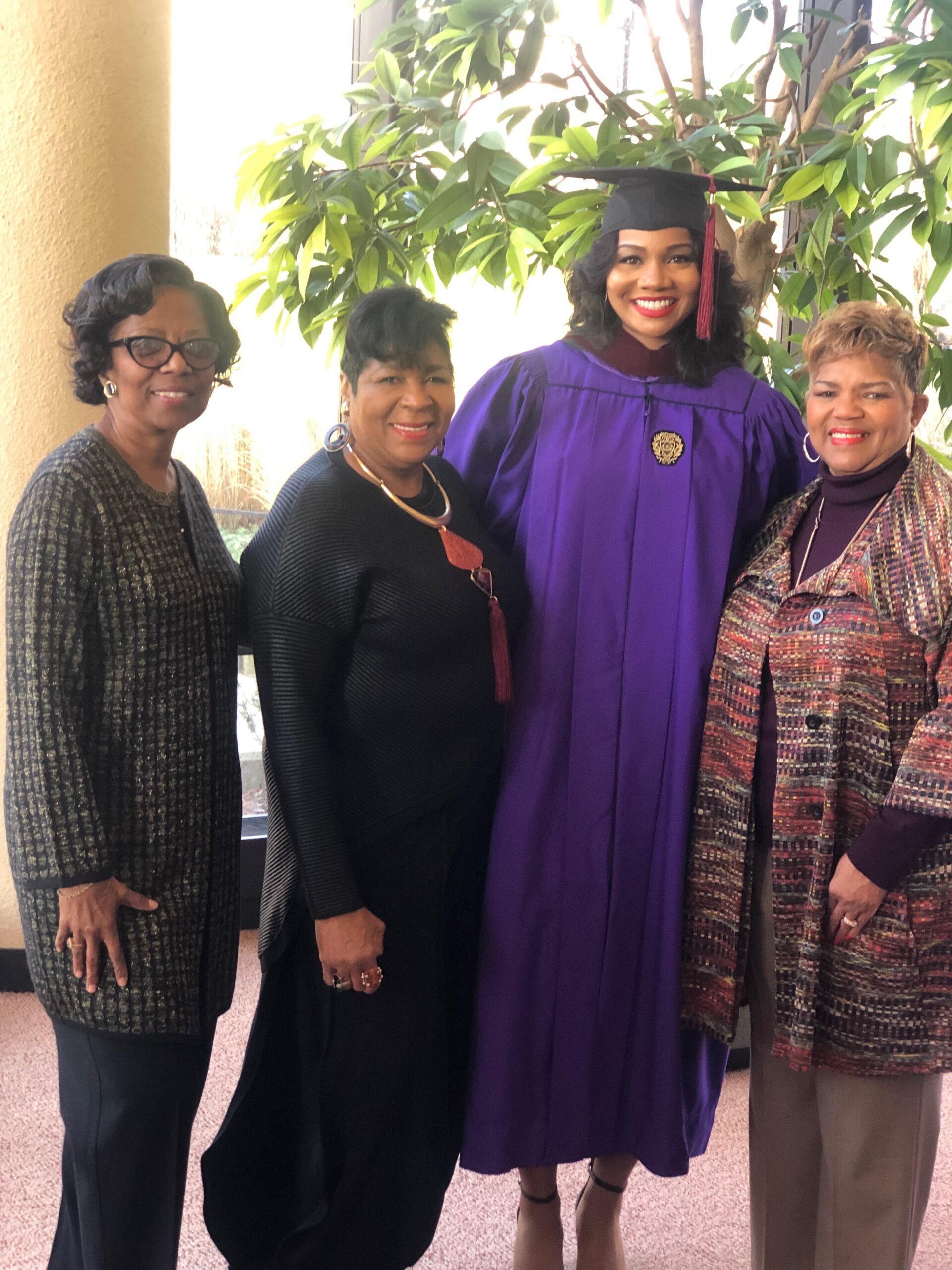 Graduate in a purple cap and gown stands with three smiling relatives indoors, leafy plants in the background.