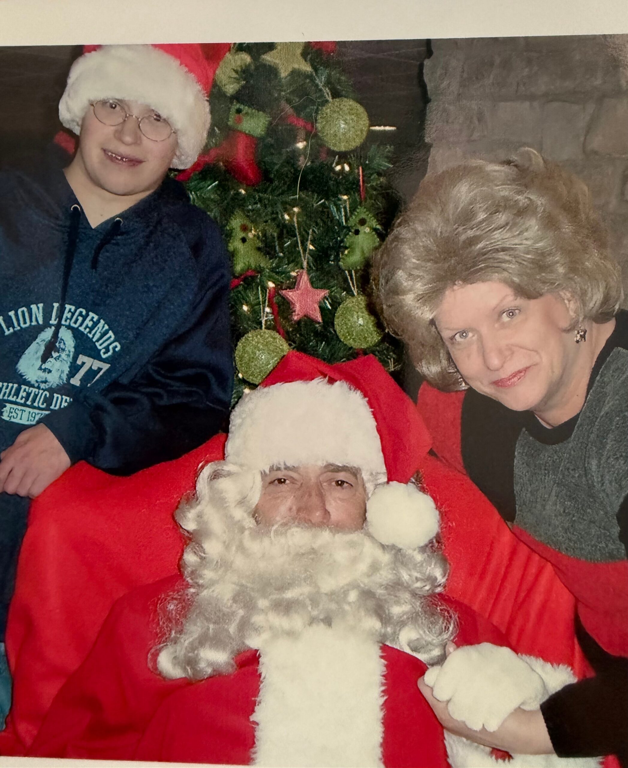 Two people posing with Santa: a man in a red Santa suit and white beard in the foreground, with a woman and a Christmas tree in the background.