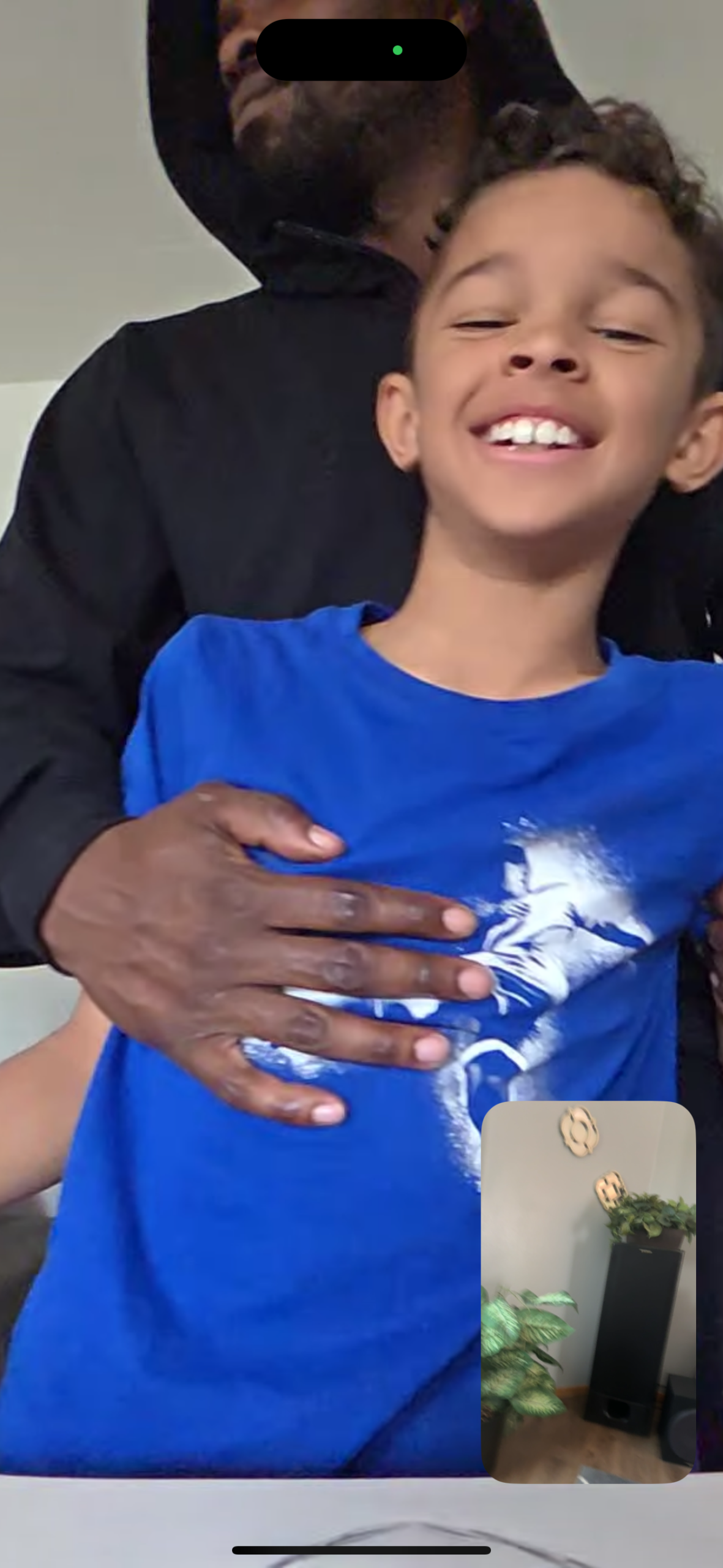 Adult man in a black hoodie hugs a smiling boy in a blue shirt from behind.