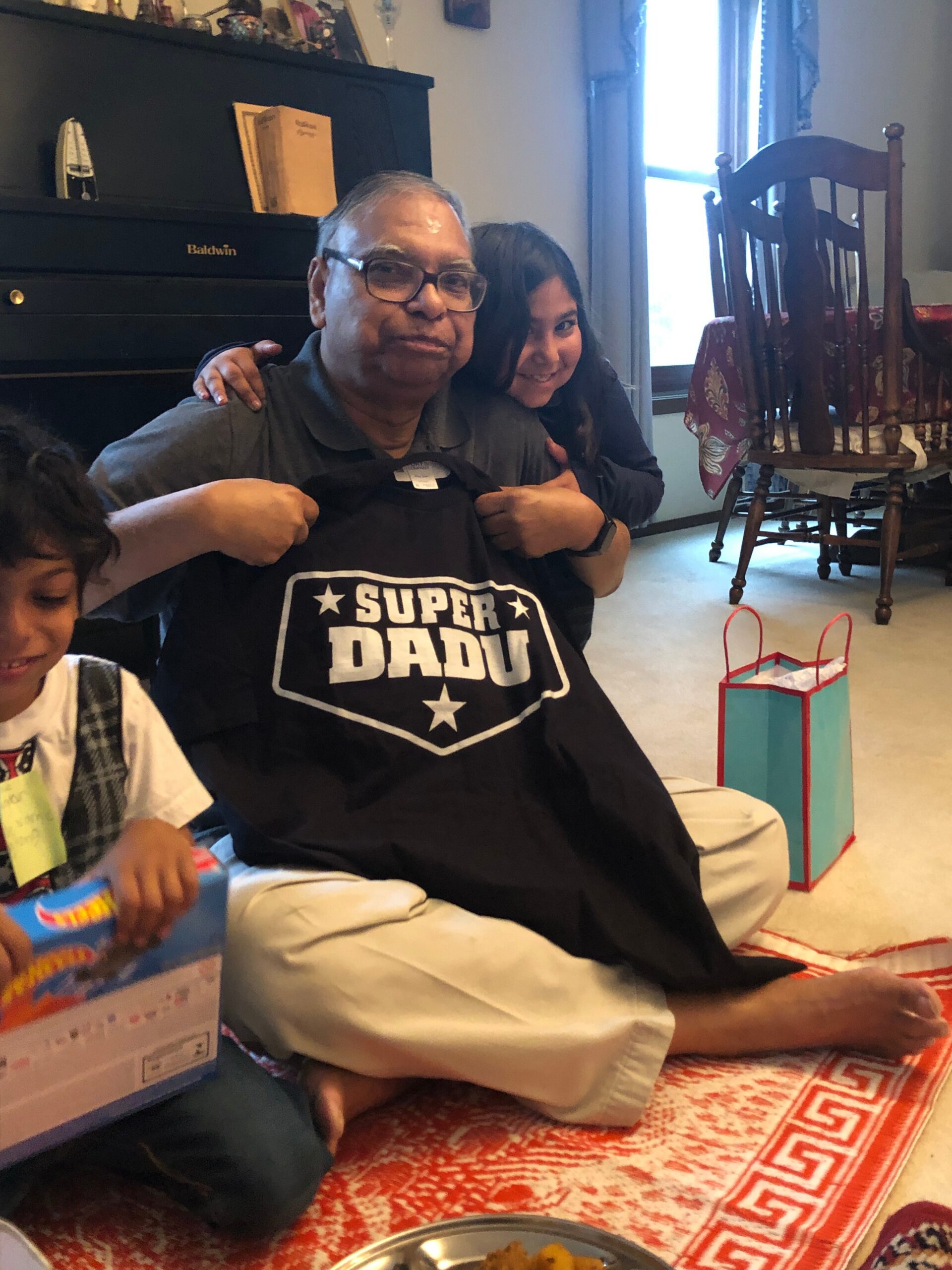 Smiling man with glasses sits on the floor as a girl hugs him from behind; he wears a black shirt that says 'SUPER DADU' at a family gathering.