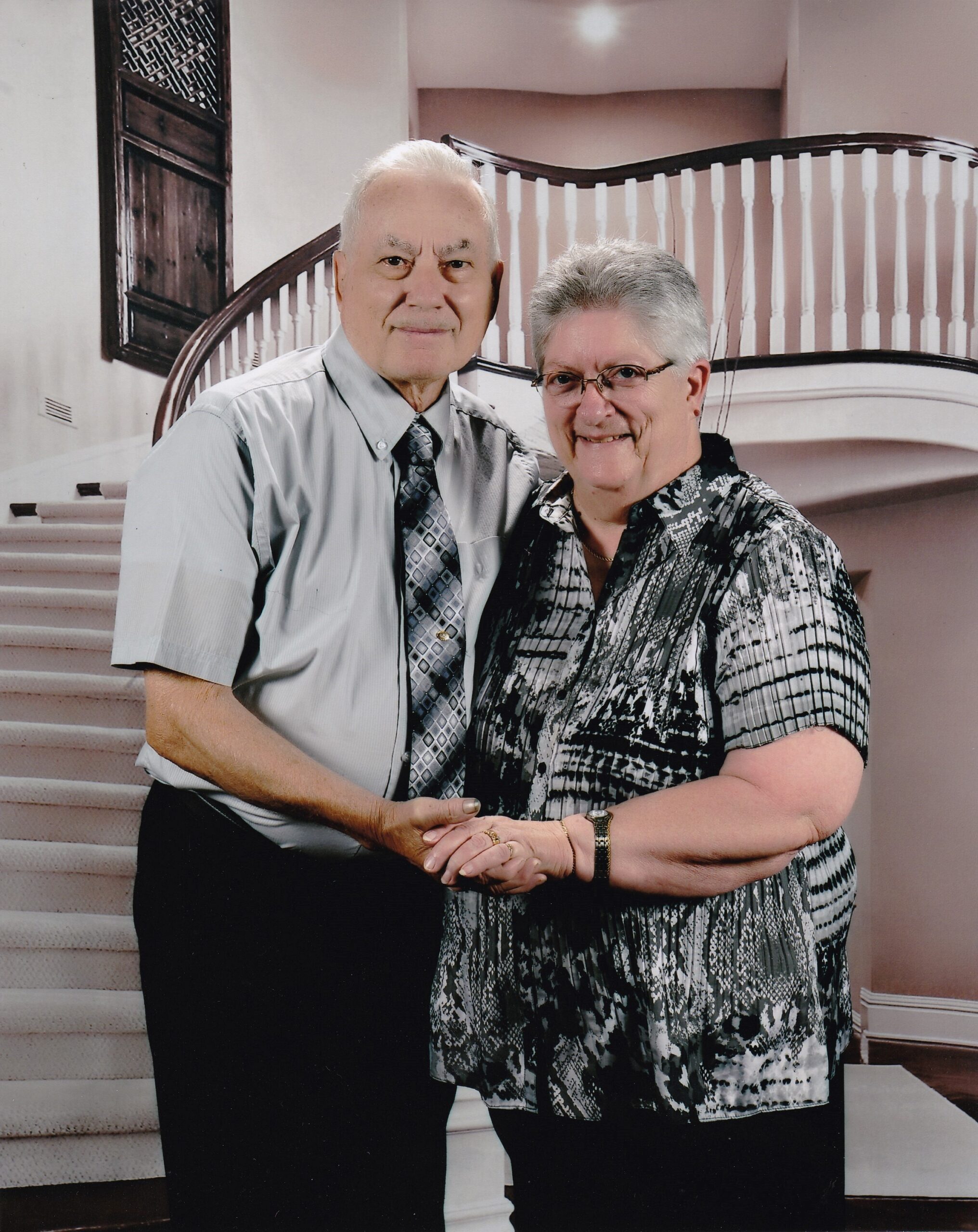 Elderly couple with arms around each other posing on a curved staircase indoors, both smiling at the camera.