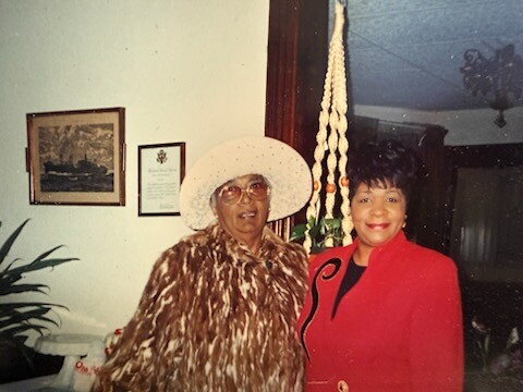 Two women posing indoors; one wearing a white sun hat and leopard fur coat, the other in a red blazer.