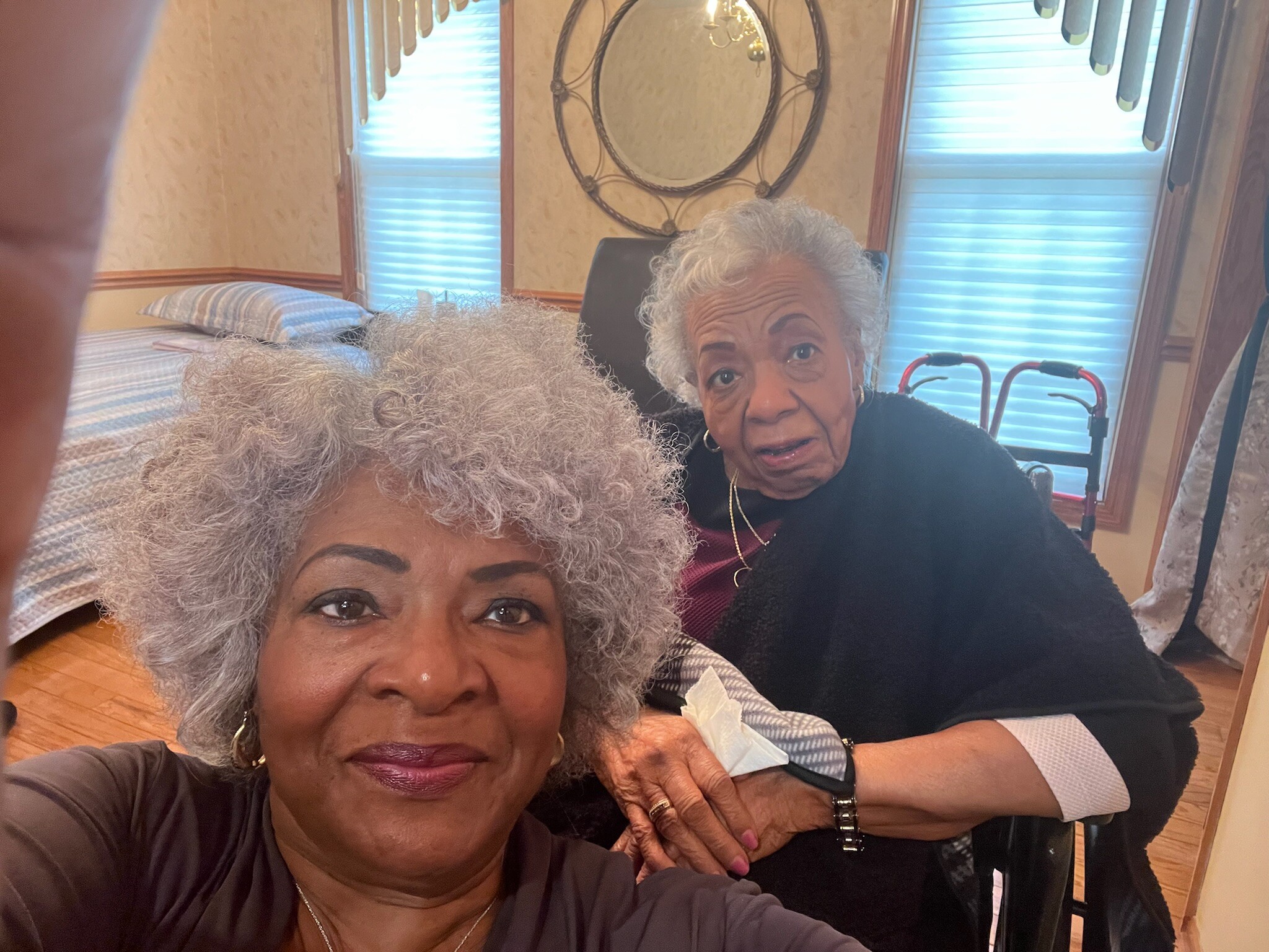 Three elderly women posing for a selfie in a cozy living room, with a walker visible in the background.