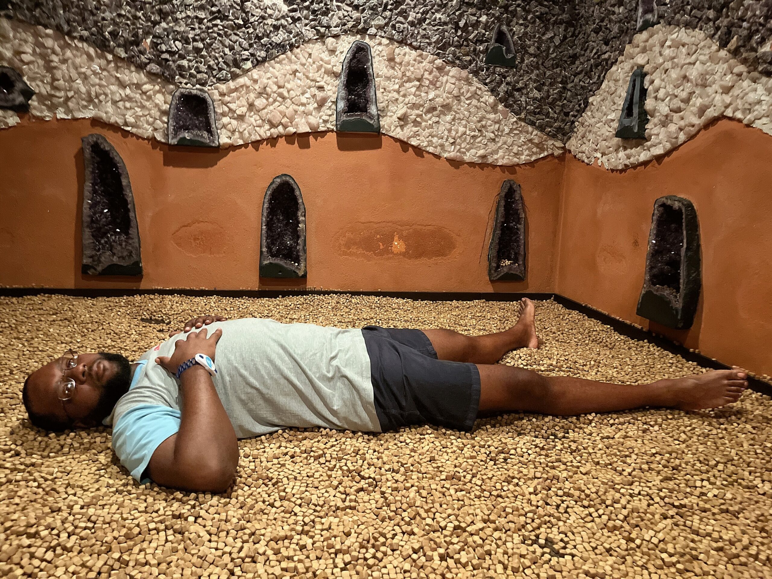 Man lying on a pebble floor in a cave-like spa room with dark stone niches along the wall behind him