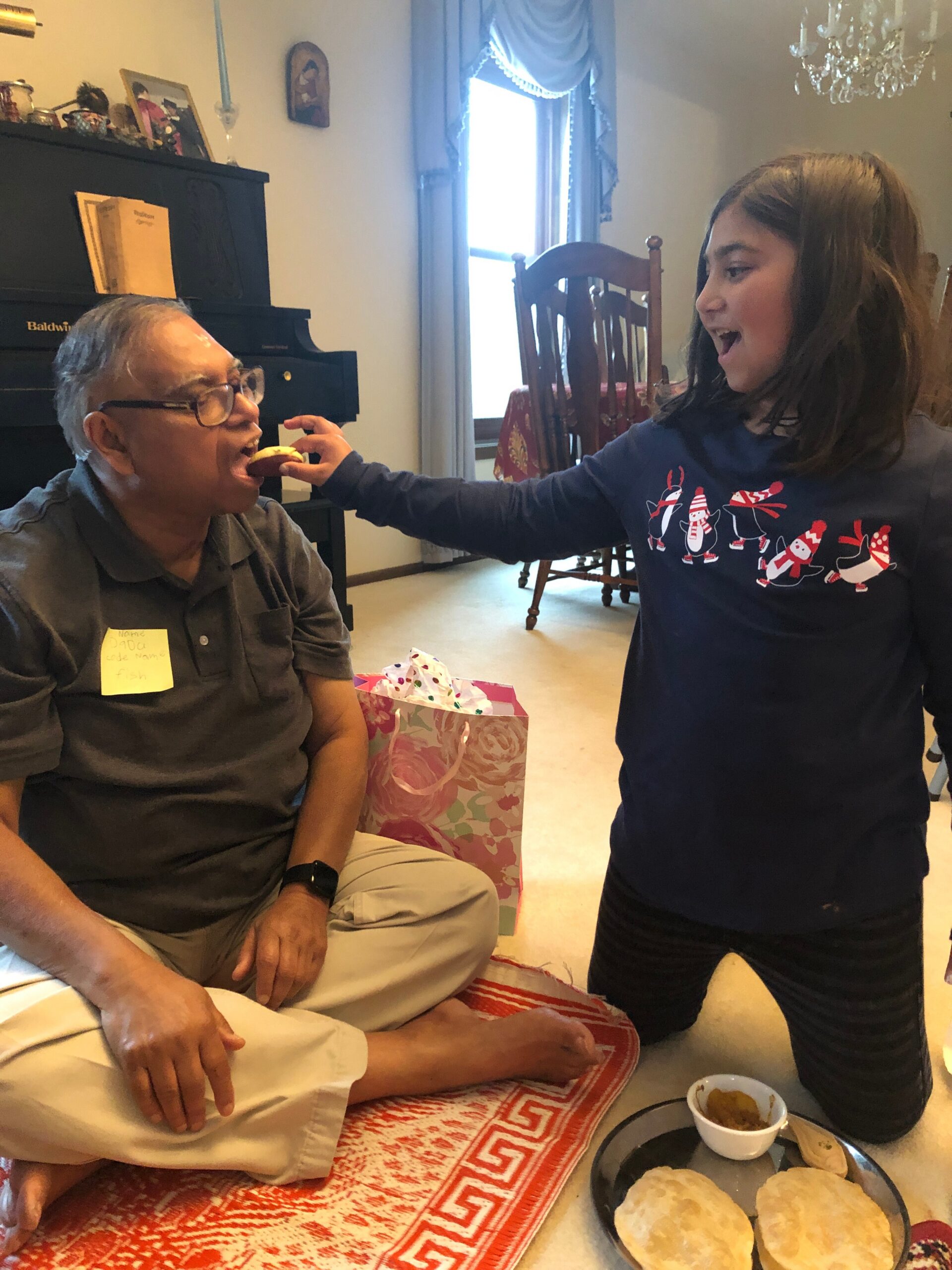 Girl feeds an elderly man a pastry while he sits on a couch beside a piano; gift bag and a tray of bread nearby.