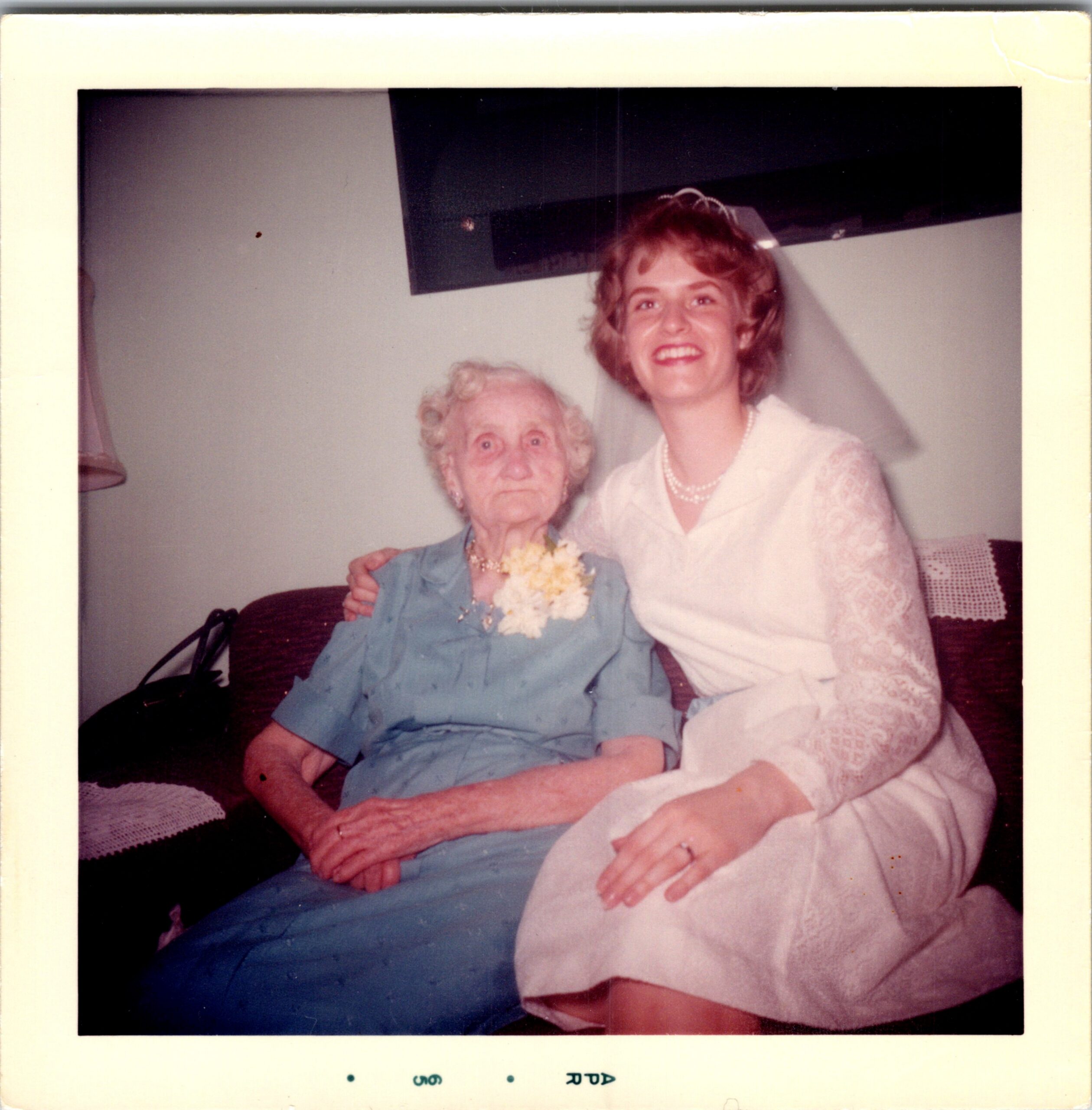 A smiling young bride in a white dress poses with an elderly woman in a blue outfit holding a bouquet at a celebration.