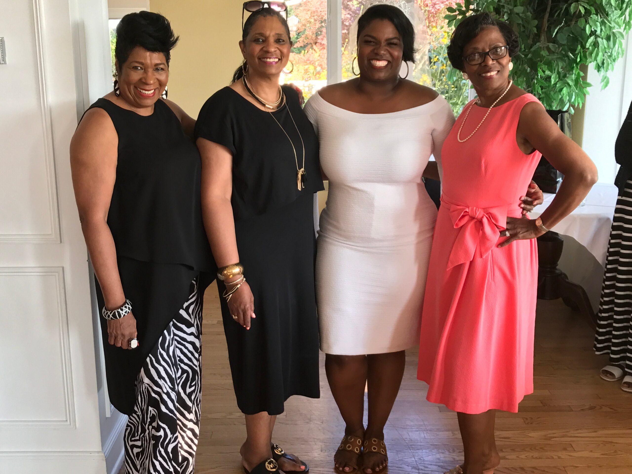 Four women in stylish dresses pose together indoors, smiling at the camera in a bright room.