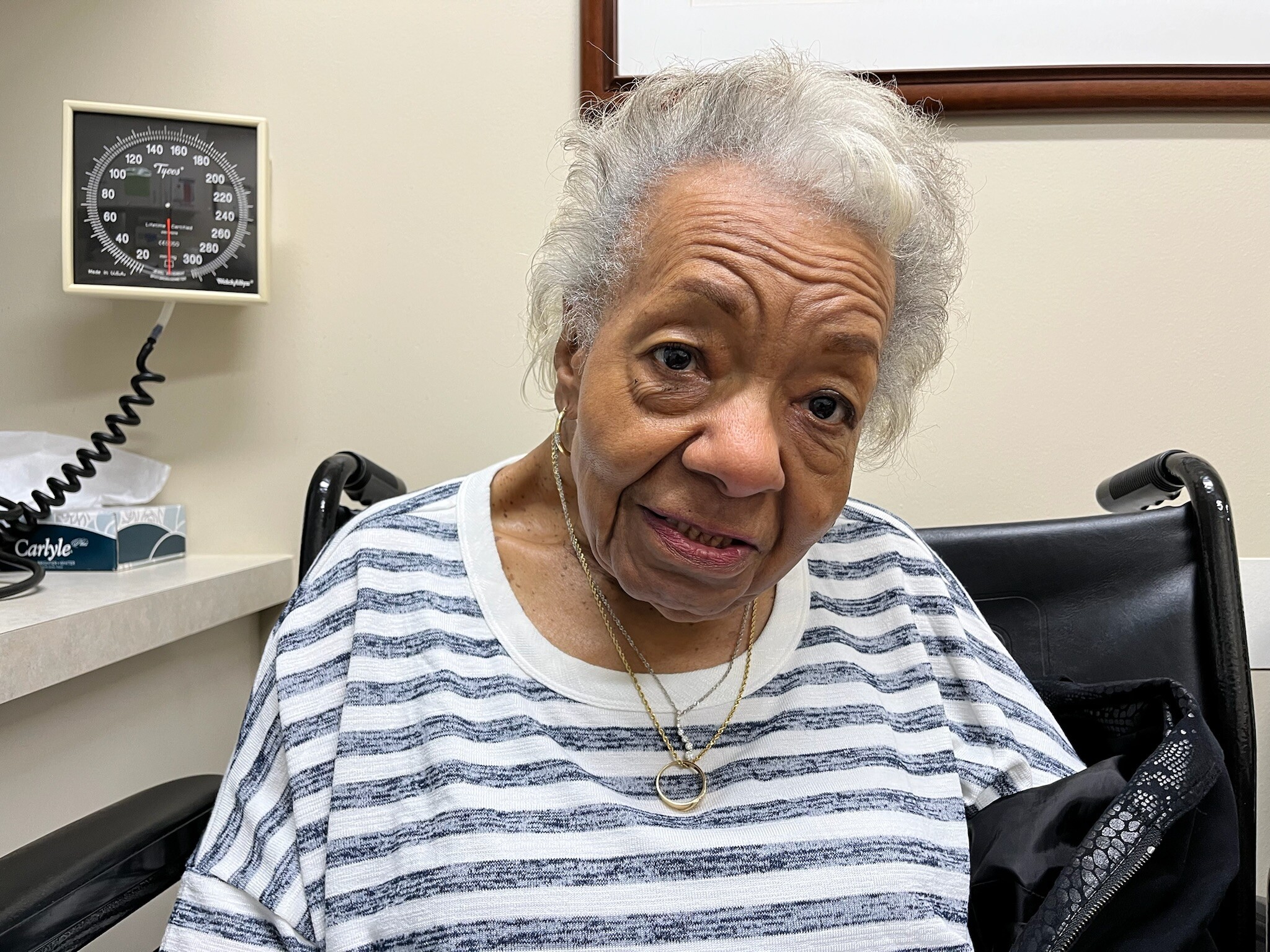 Older woman in a clinic with a blood pressure cuff on her arm and a wall gauge in the background.