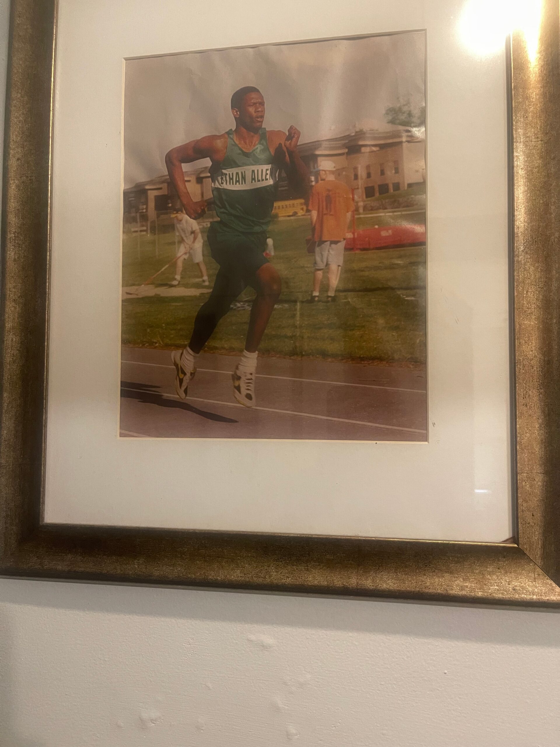 Framed photo of a male sprinter in a green uniform running on a track, bib reads NATHAN ALLEN, with onlookers and campus buildings in the background.
