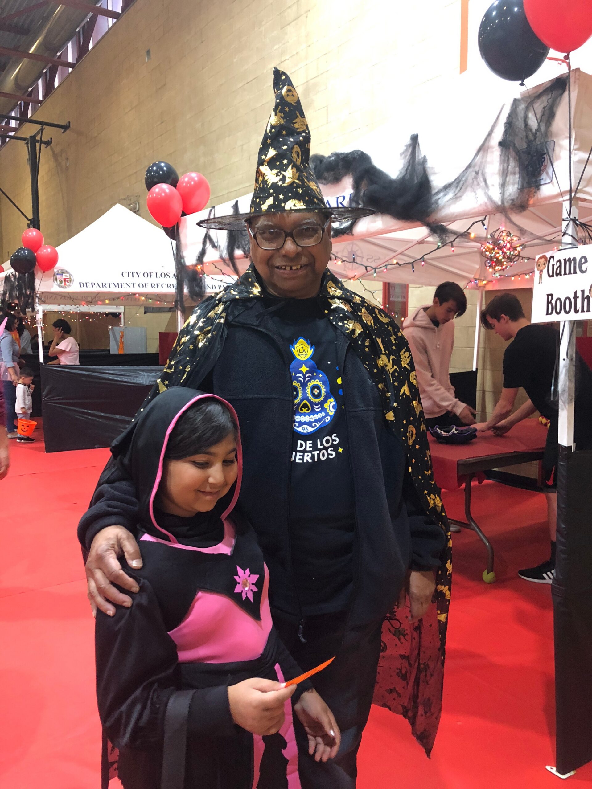 Smiling man in a black and gold starry wizard cape and tall pointy hat posing with a child at a Halloween booth. Behind them are tents, balloons, and festive decorations.