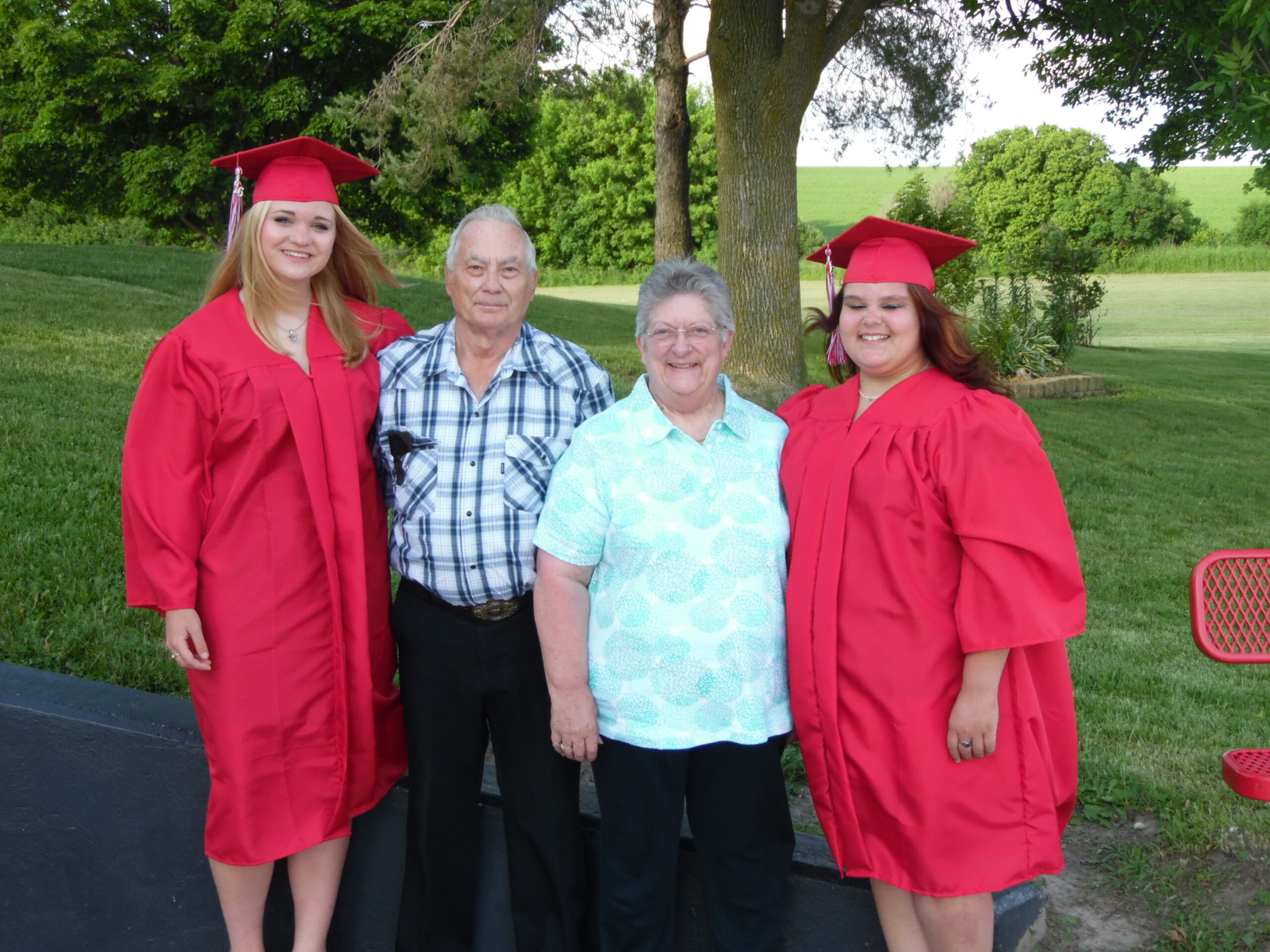 Four people pose outdoors for a graduation photo: two young women in red gowns and mortarboards flanking an older couple under trees in a green park.