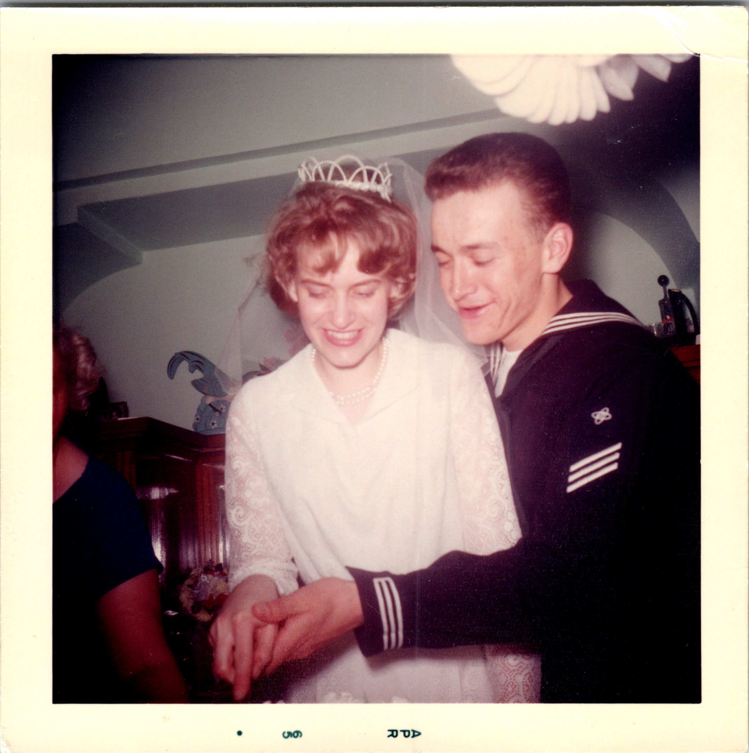 Bride wearing tiara and veil, pearl necklace, smiling as she dances with a man in a dark naval uniform at a wedding reception.