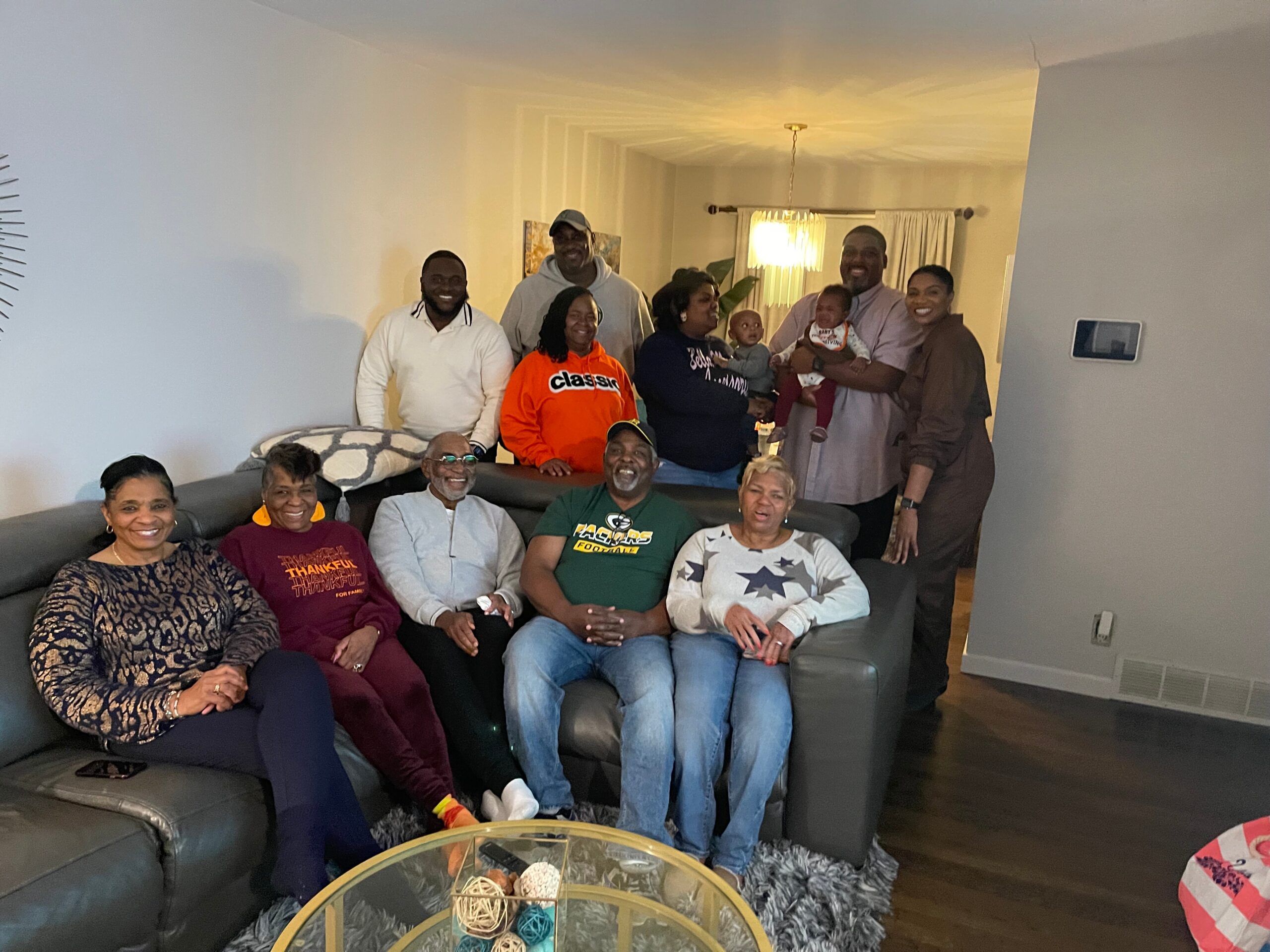 A multi generational family gathers in a living room, posing for a group photo with smiles and relaxed seating on a sofa.