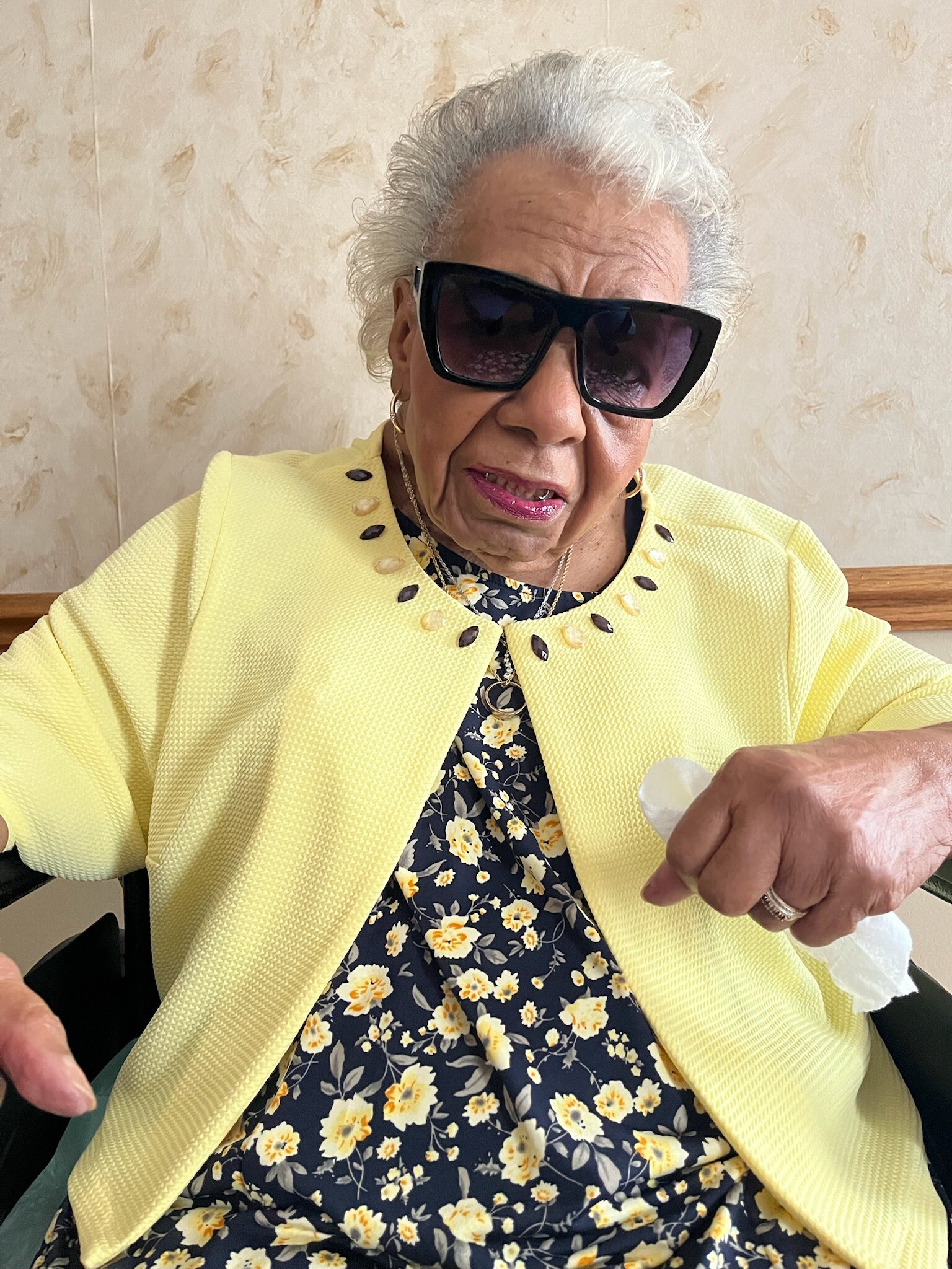 Older woman with white hair wearing oversized black sunglasses and a bright yellow textured jacket over a navy floral dress indoors.