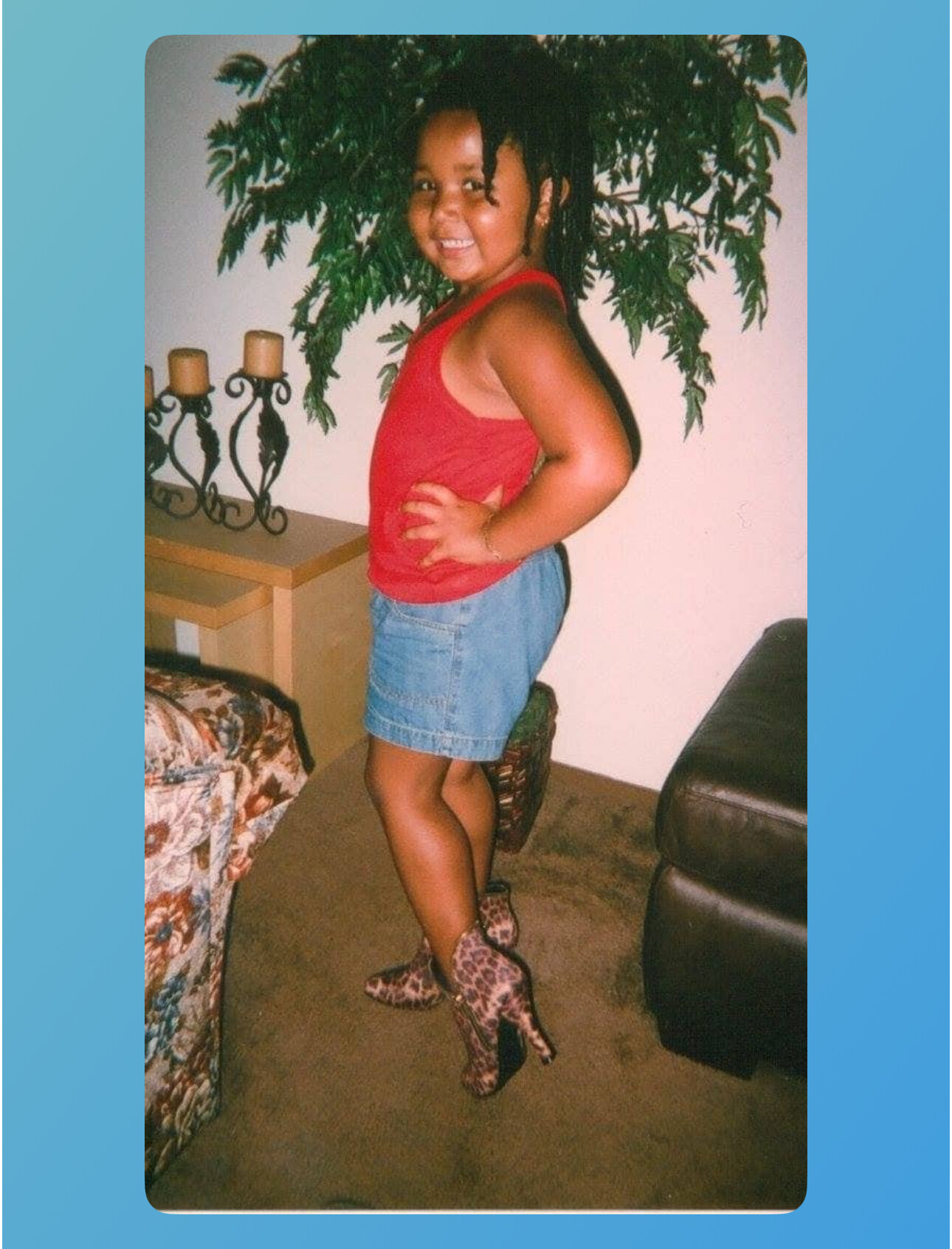 Young child posing in a red tank top, denim shorts, and leopard-print boots indoors with a large plant behind.