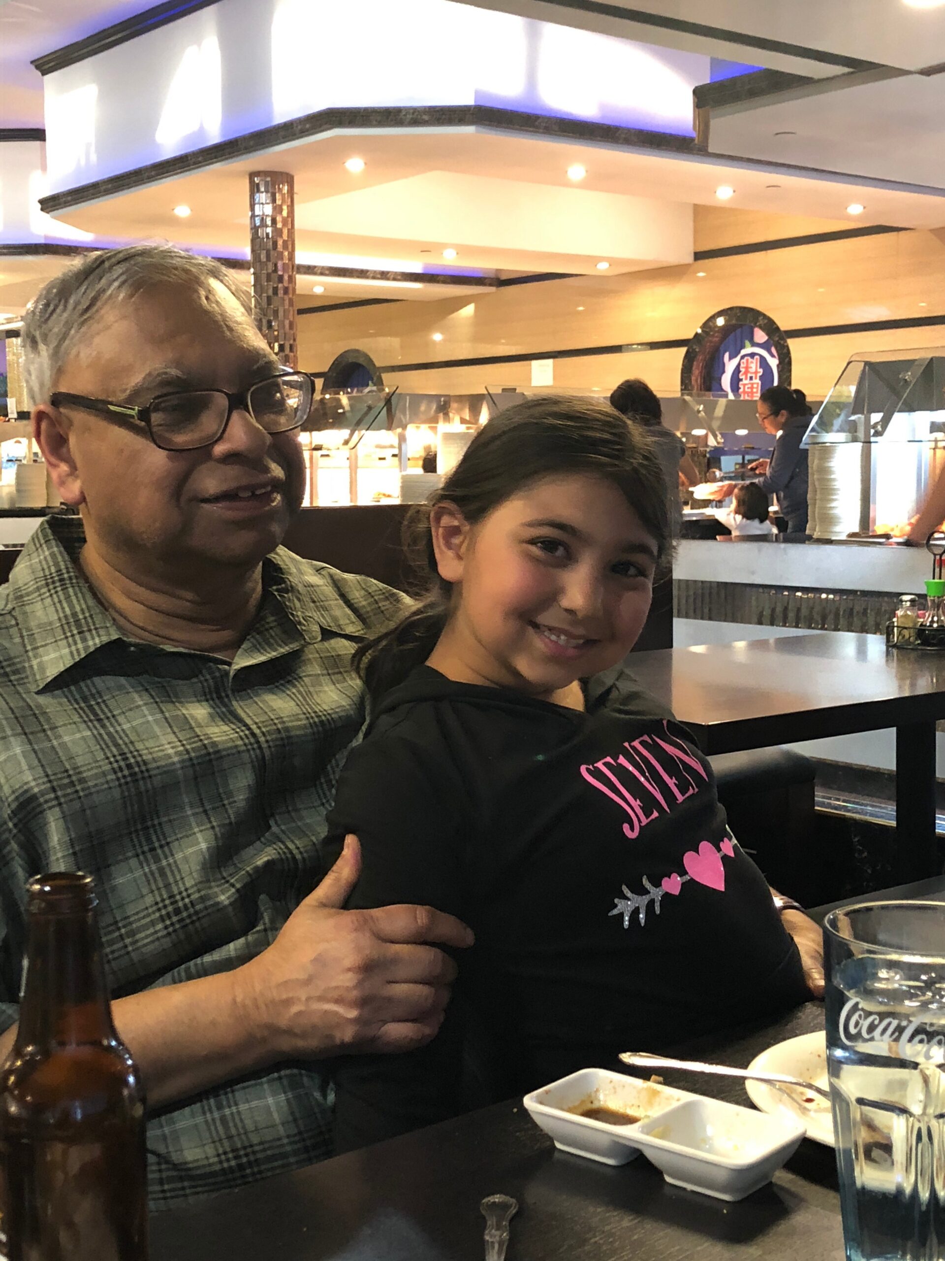 Older man with glasses and a smiling young girl at a restaurant table, with dipping sauces and a Coca-Cola glass in view, buffet area in the background.