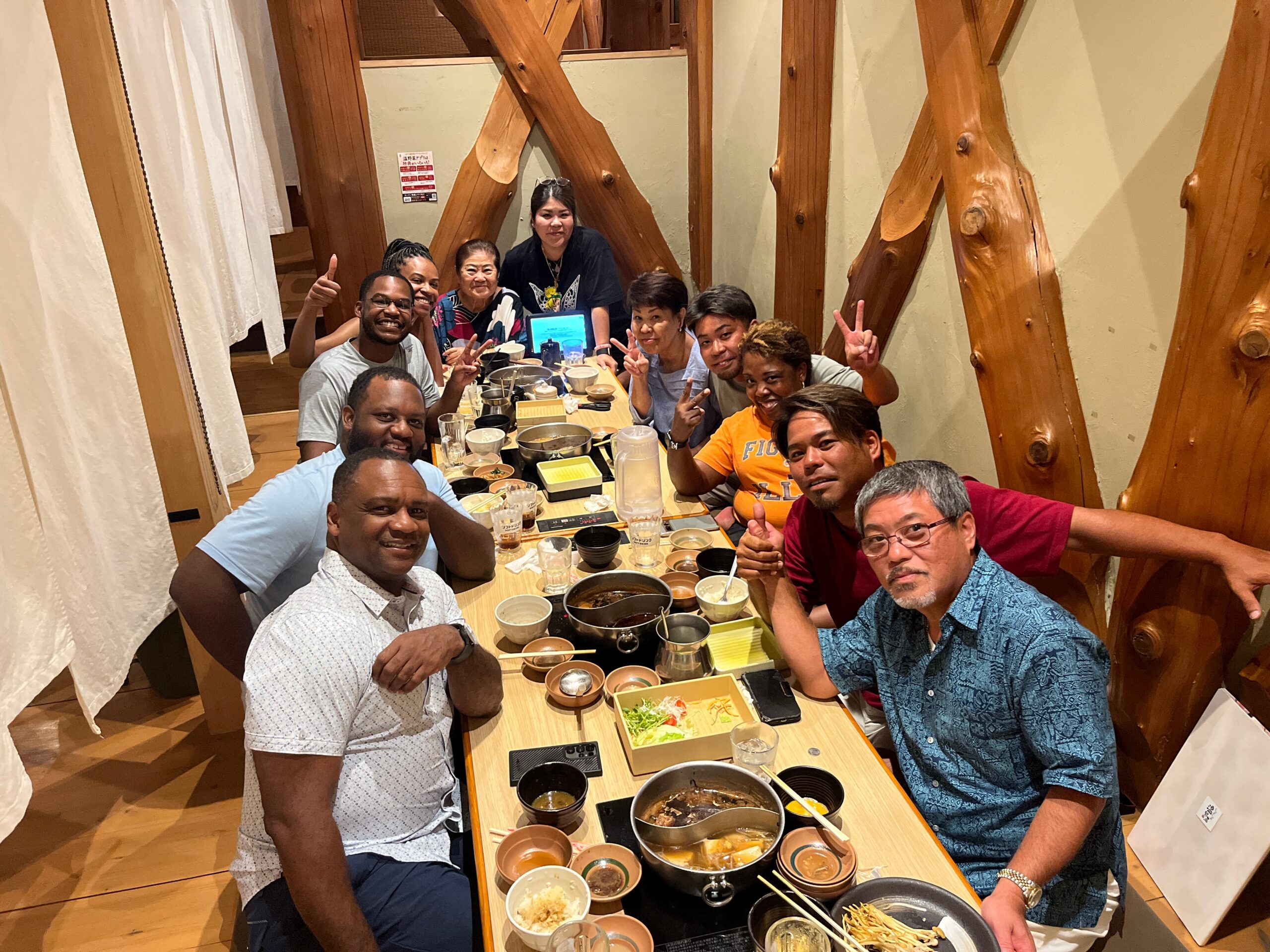 A diverse group of friends dining together at a long wooden table in a cozy restaurant, posing and smiling for the photo.
