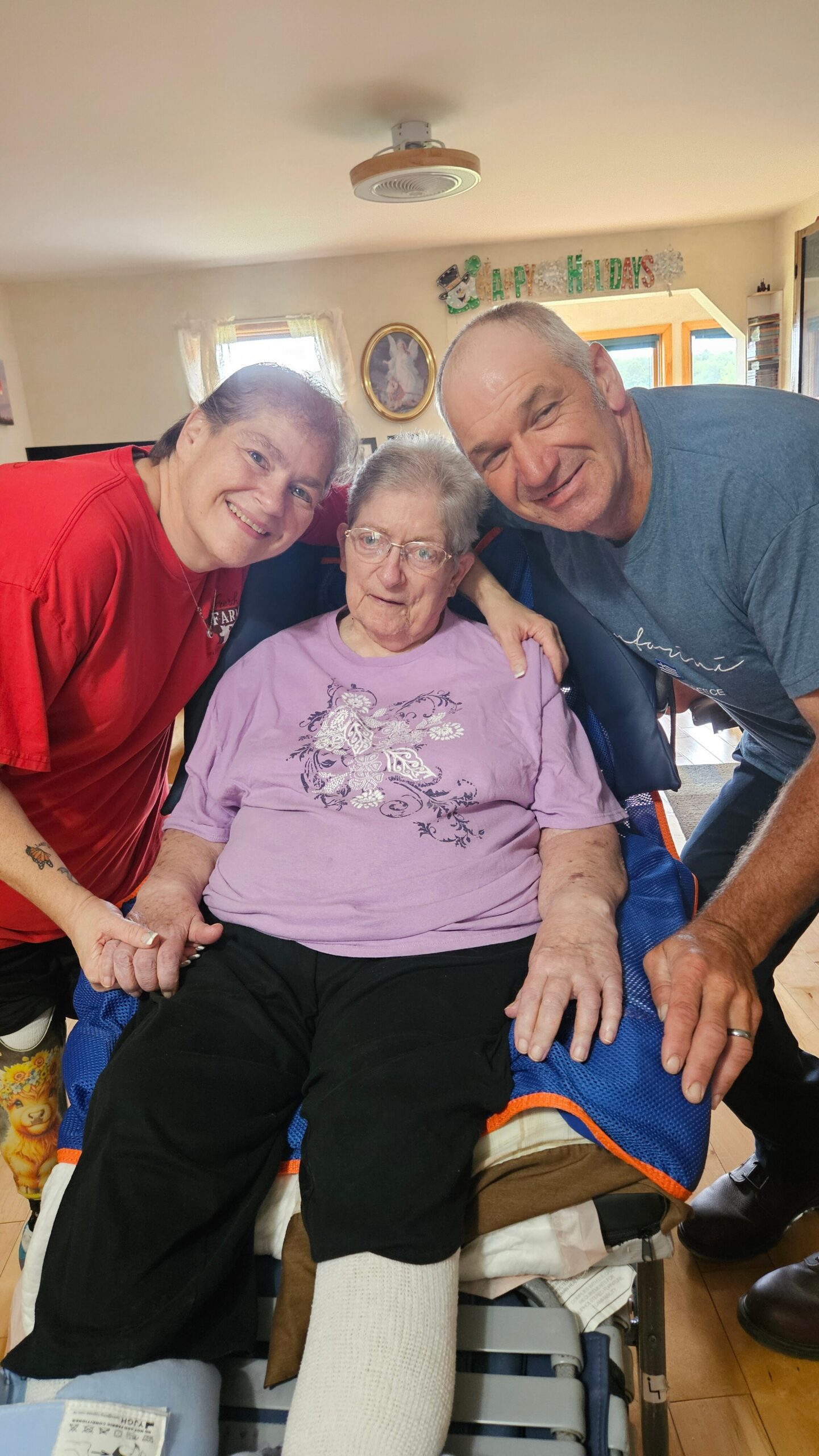 Three people smiling for a photo: an elderly woman in a purple shirt seated in a wheelchair, flanked by a woman in red and a man in blue, indoors with a 'Holidays' banner in the background.