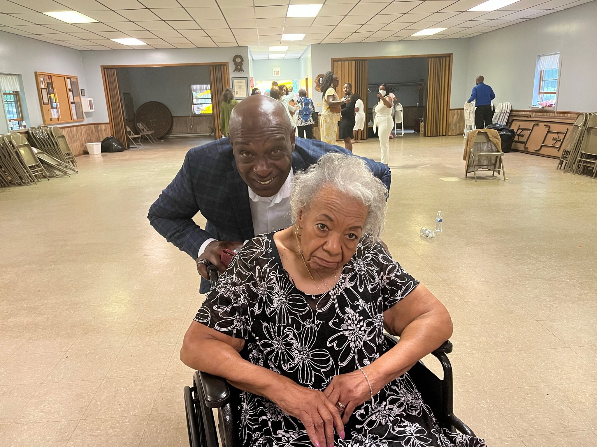 Elderly woman in a floral dress sits in a wheelchair at the foreground, with a smiling man leaning over from behind in a large event hall.