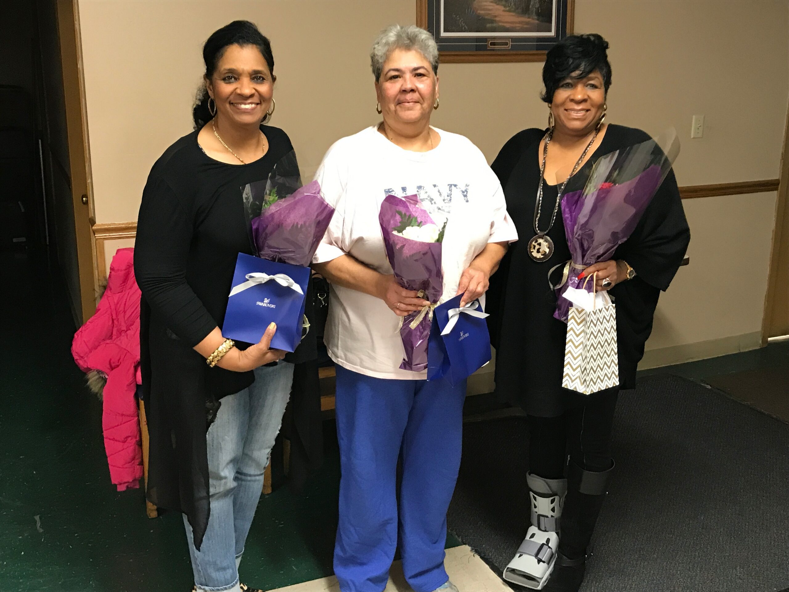 Three women indoors, smiling; center woman holds a purple bouquet, others carry blue gift bags.