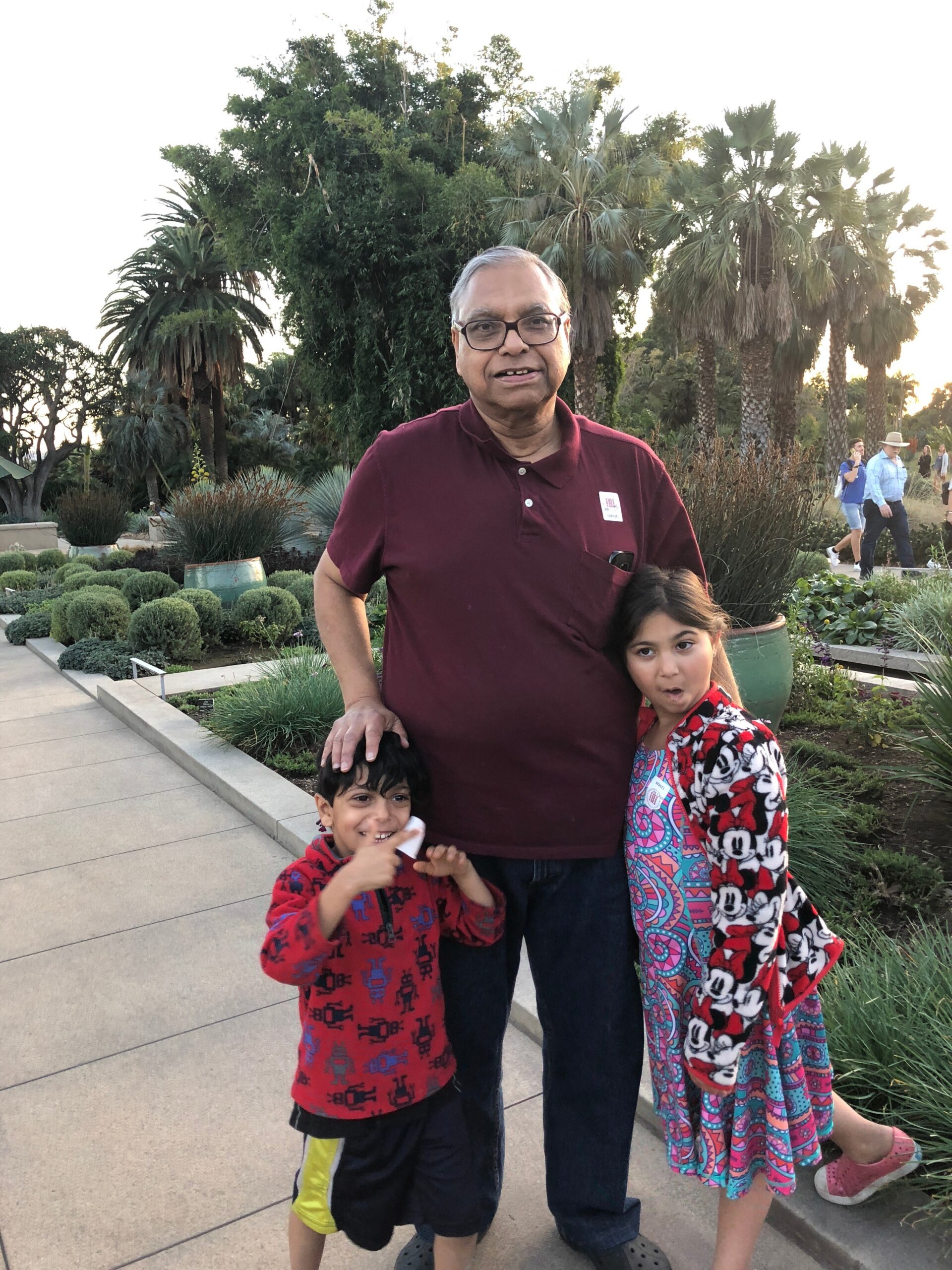 Man in maroon polo shirt with glasses posing with two children in a park; greenery and palm trees in the background.