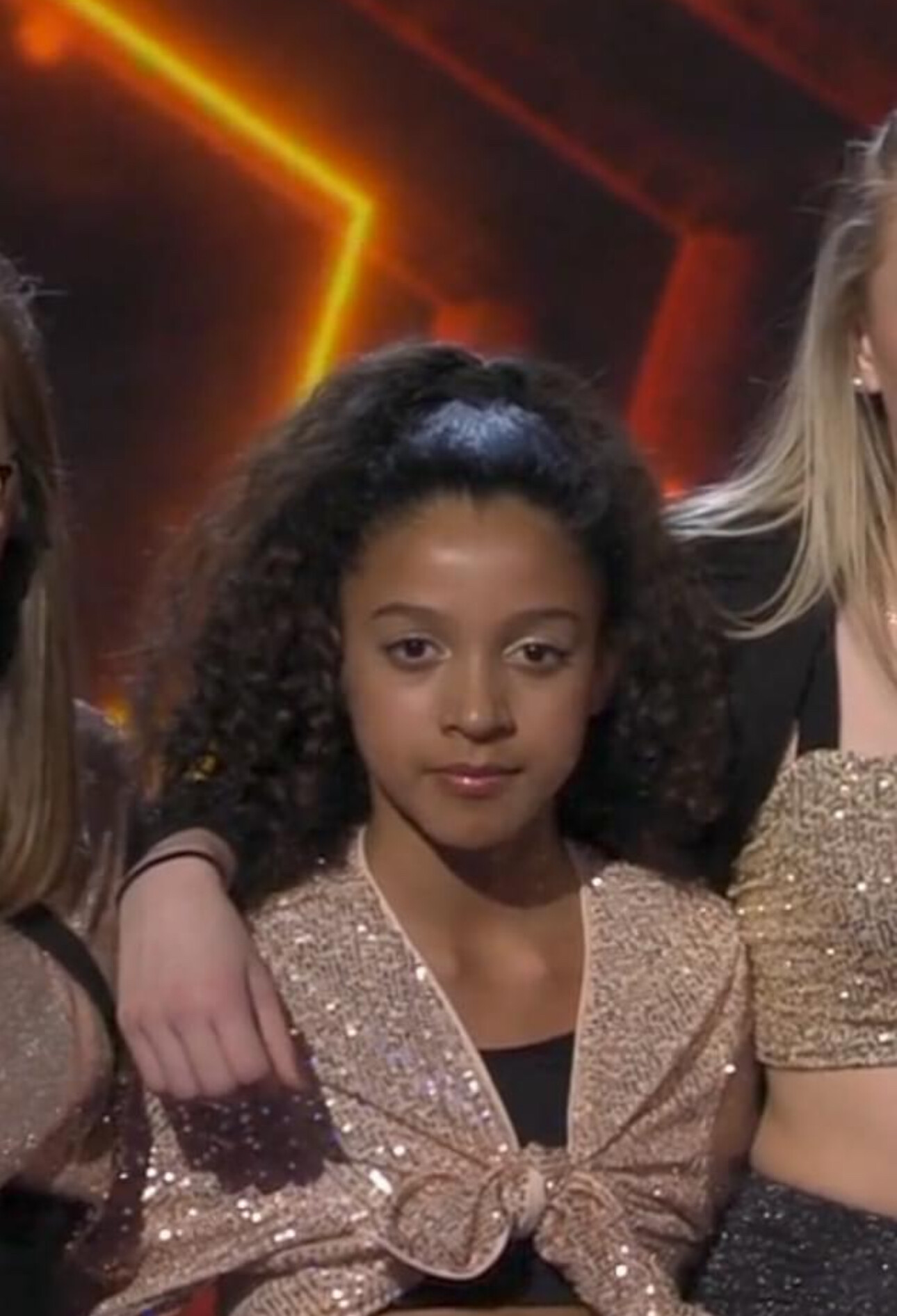 Young girl with curly hair wearing a glittery gold jacket, posing with others at a group event.