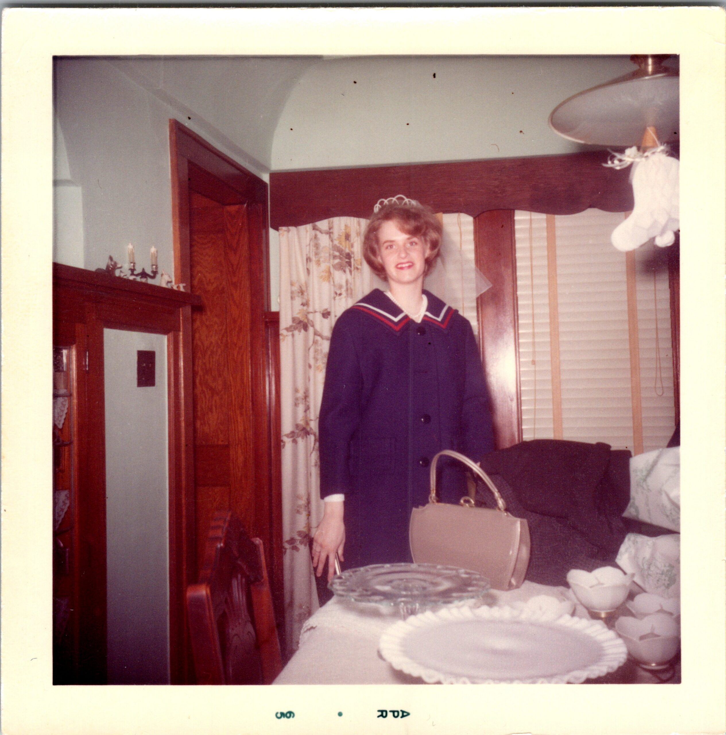 Young woman in a dark dress with white trim and a small tiara stands by a table with a decorated cake in a home dining room.