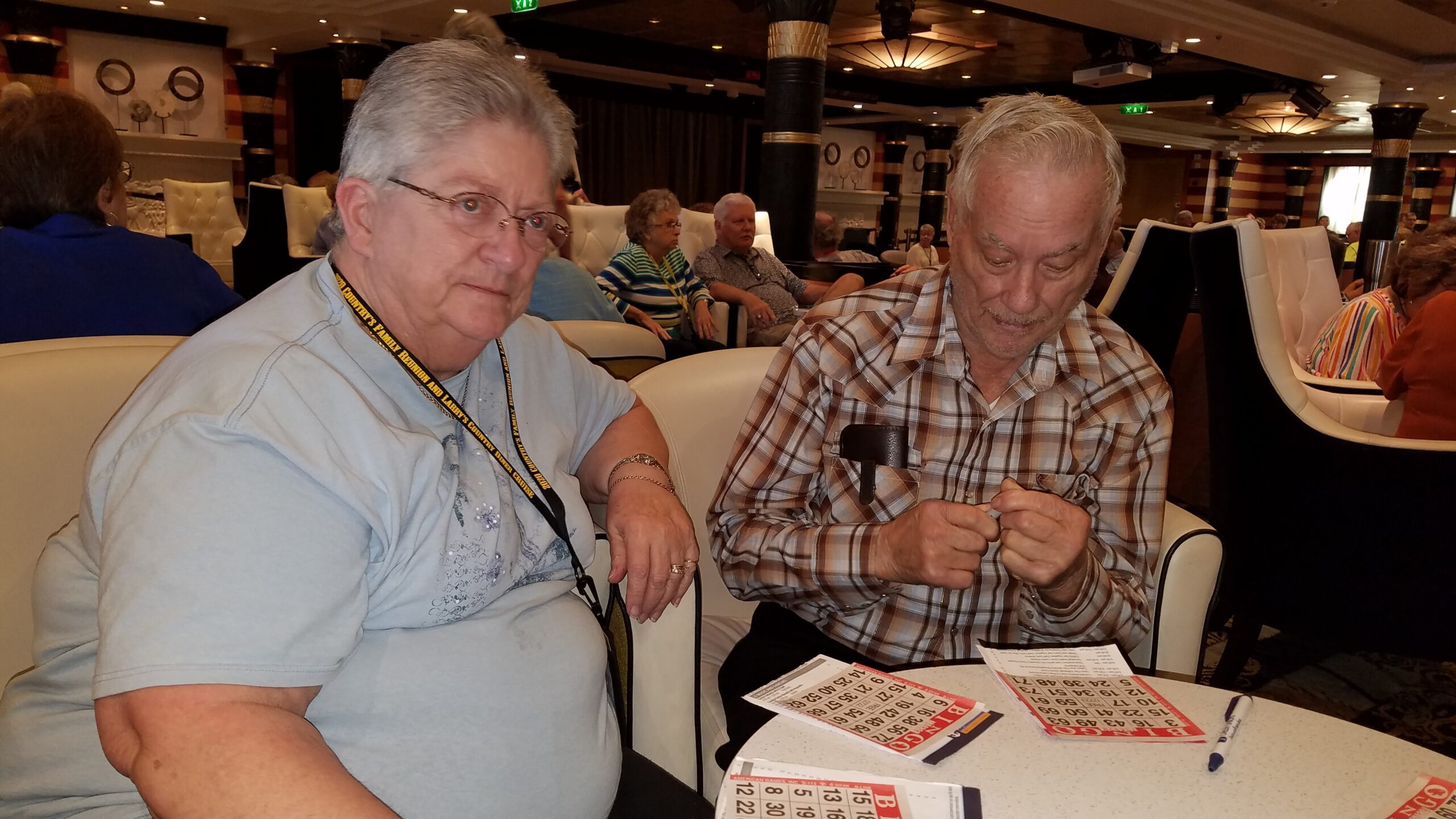 Two seniors sit at a bingo table, looking at bingo cards and markers in a busy lounge with other players in the background.