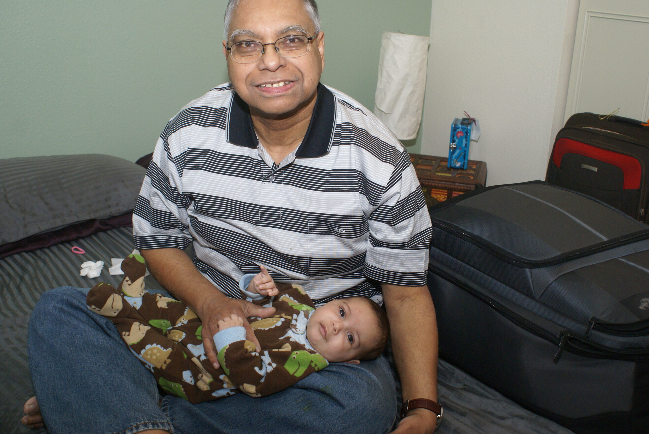 Older man with glasses sits on a bed, smiling, while holding a baby wrapped in a brown monkey-blanket.