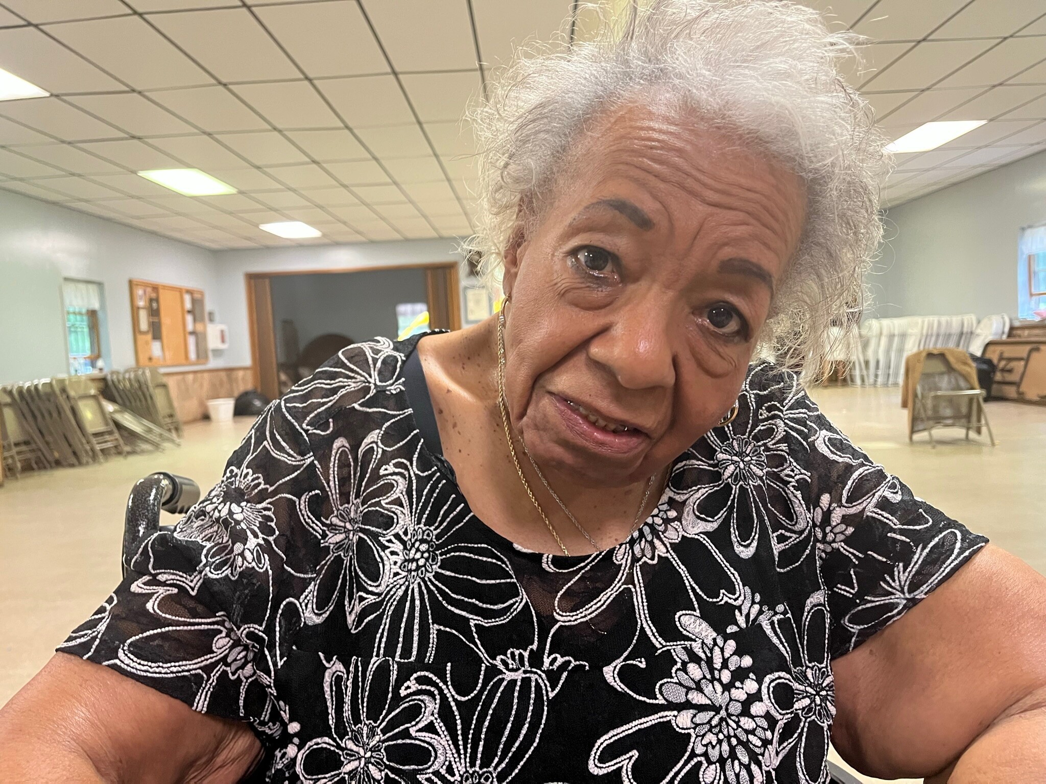 Close-up of smiling elderly woman with gray hair wearing a black and white floral blouse, taking a selfie in a room with chairs.