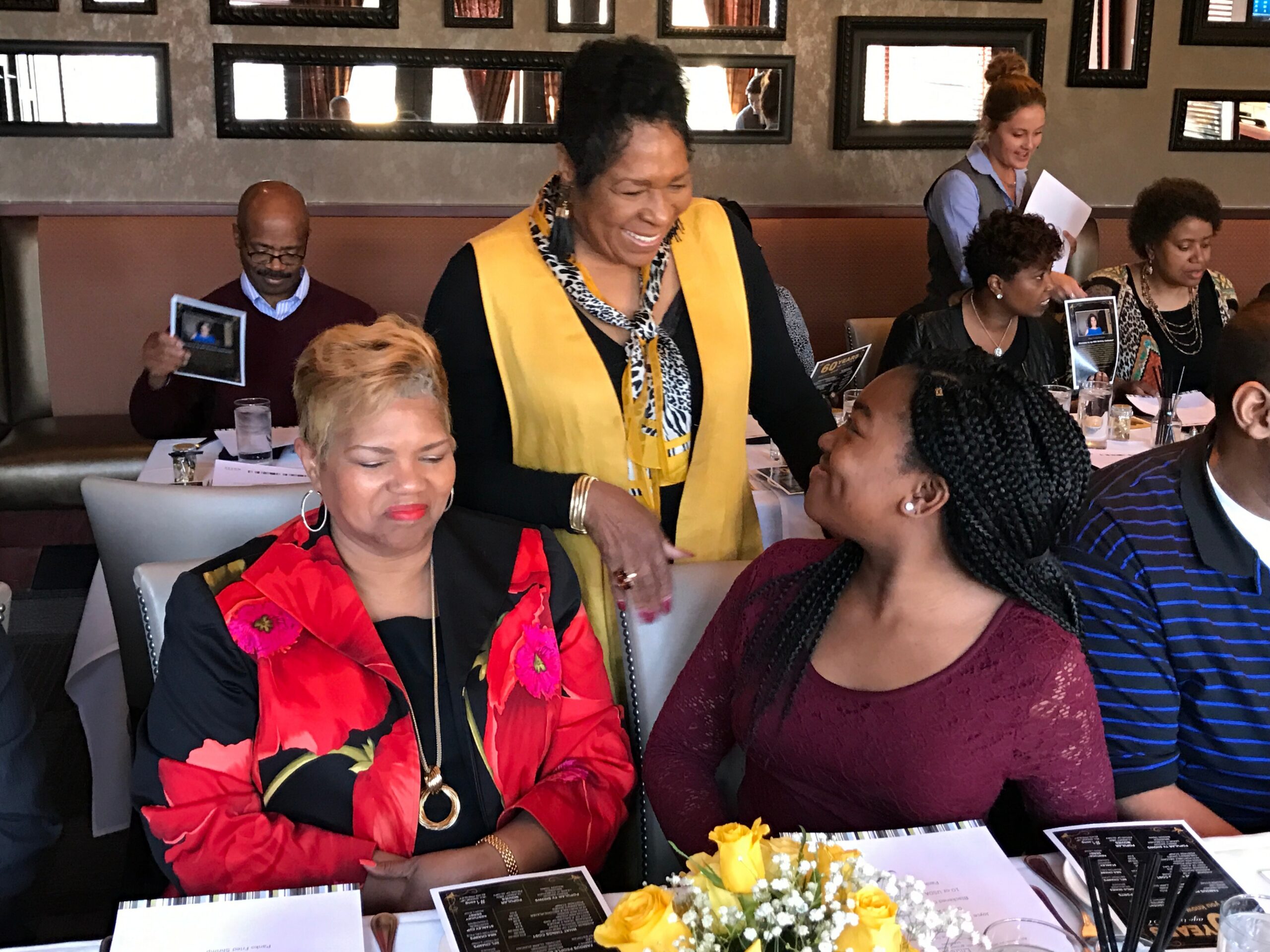 Group of people dining at a restaurant, a woman in a yellow scarf stands smiling while others sit with menus nearby.