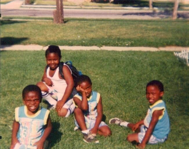 Four young children sitting on a grassy park lawn, smiling at the camera; wearing light blue and white sports outfits.