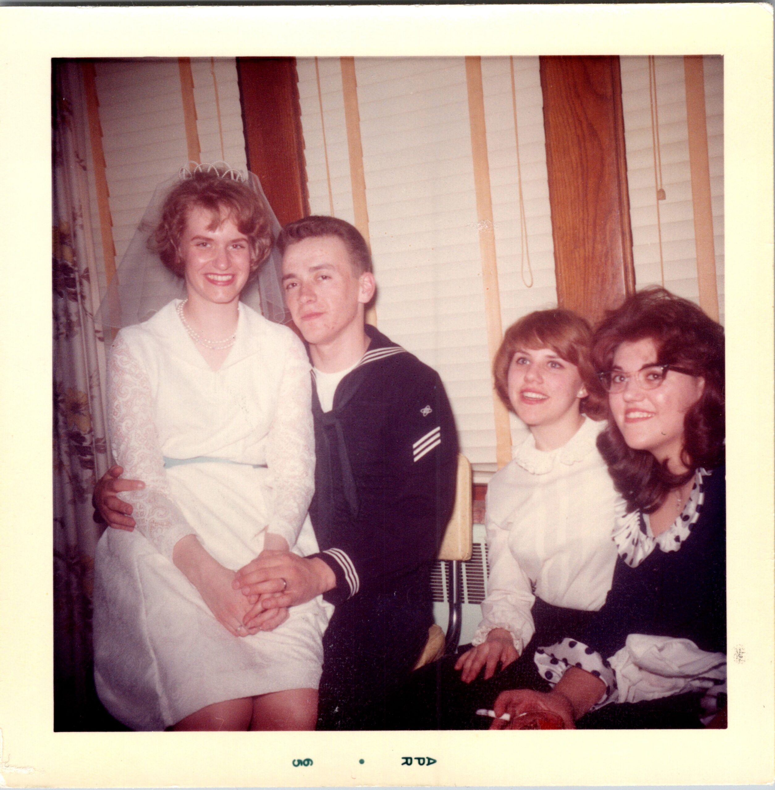 Group wedding photo: bride in a white dress with a veil stands beside a man in a navy sailor uniform, flanked by smiling guests indoors.