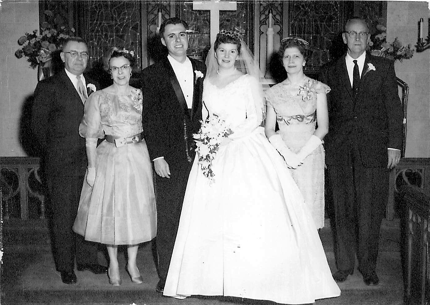 Bride and groom stand in the center with two women and two men on a church wedding backdrop, all in formal attire in a black-and-white group portrait.