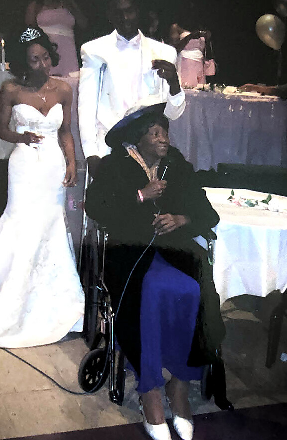 Elderly woman in a wheelchair wearing a hat and blue skirt, speaking into a microphone at a wedding reception, with a bride in a white gown in the background.