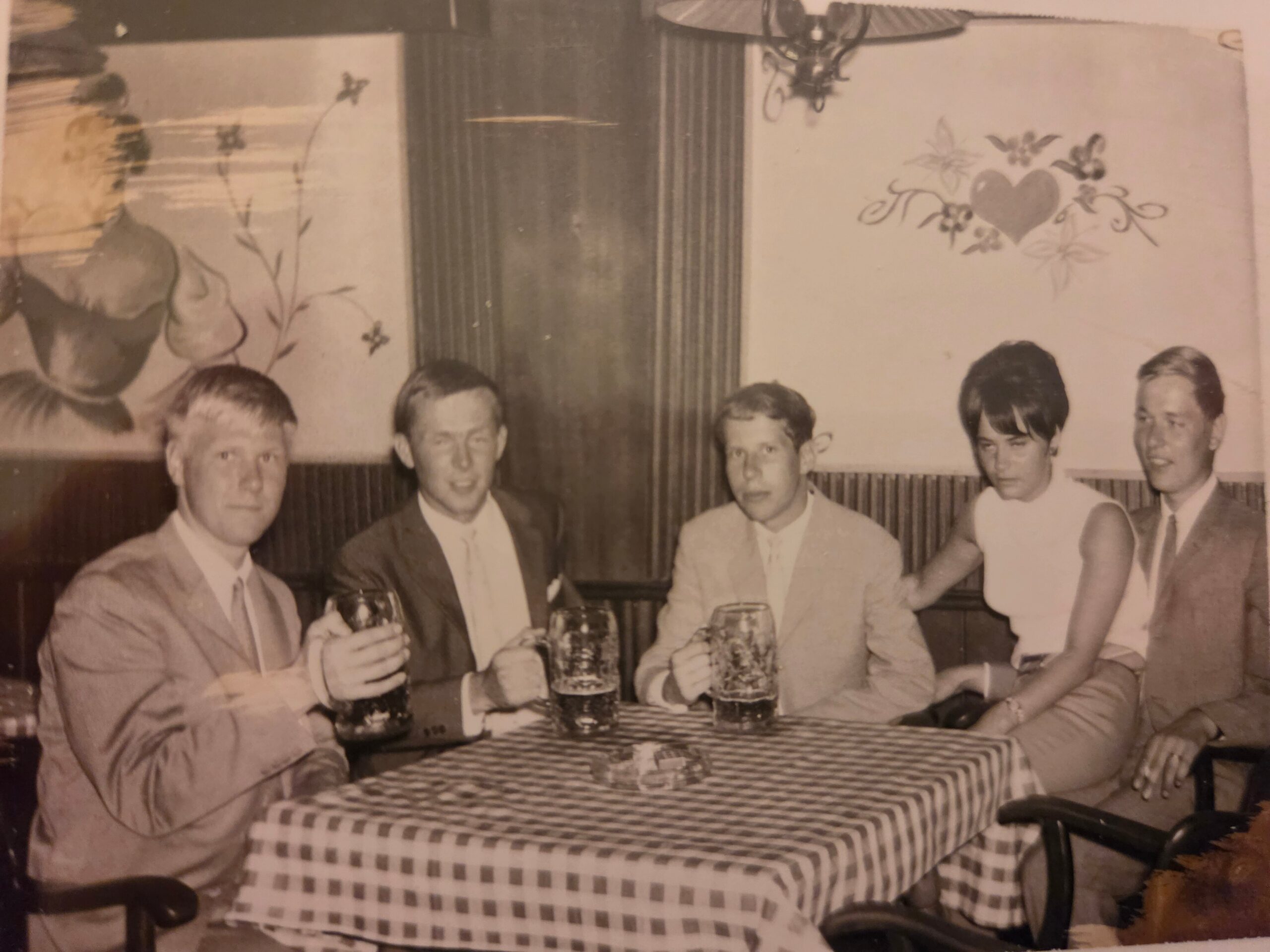 Five men in suits sit at a checked-tablecloth restaurant table, toasting with beer mugs in a vintage photo.