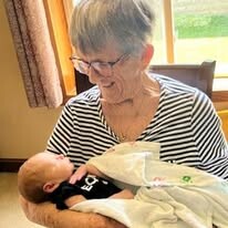 Older woman with glasses smiles as she cradles a newborn baby wrapped in a light blanket.