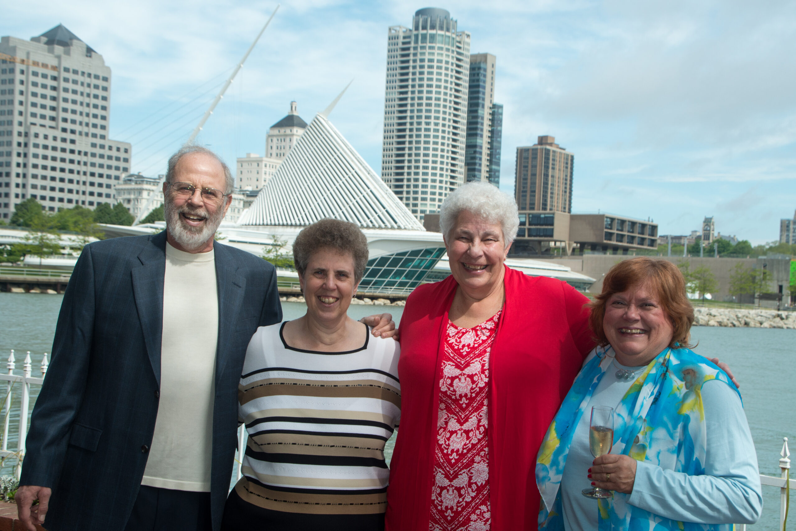 Group of four smiling adults posing together by a waterfront with modern city buildings in the background.