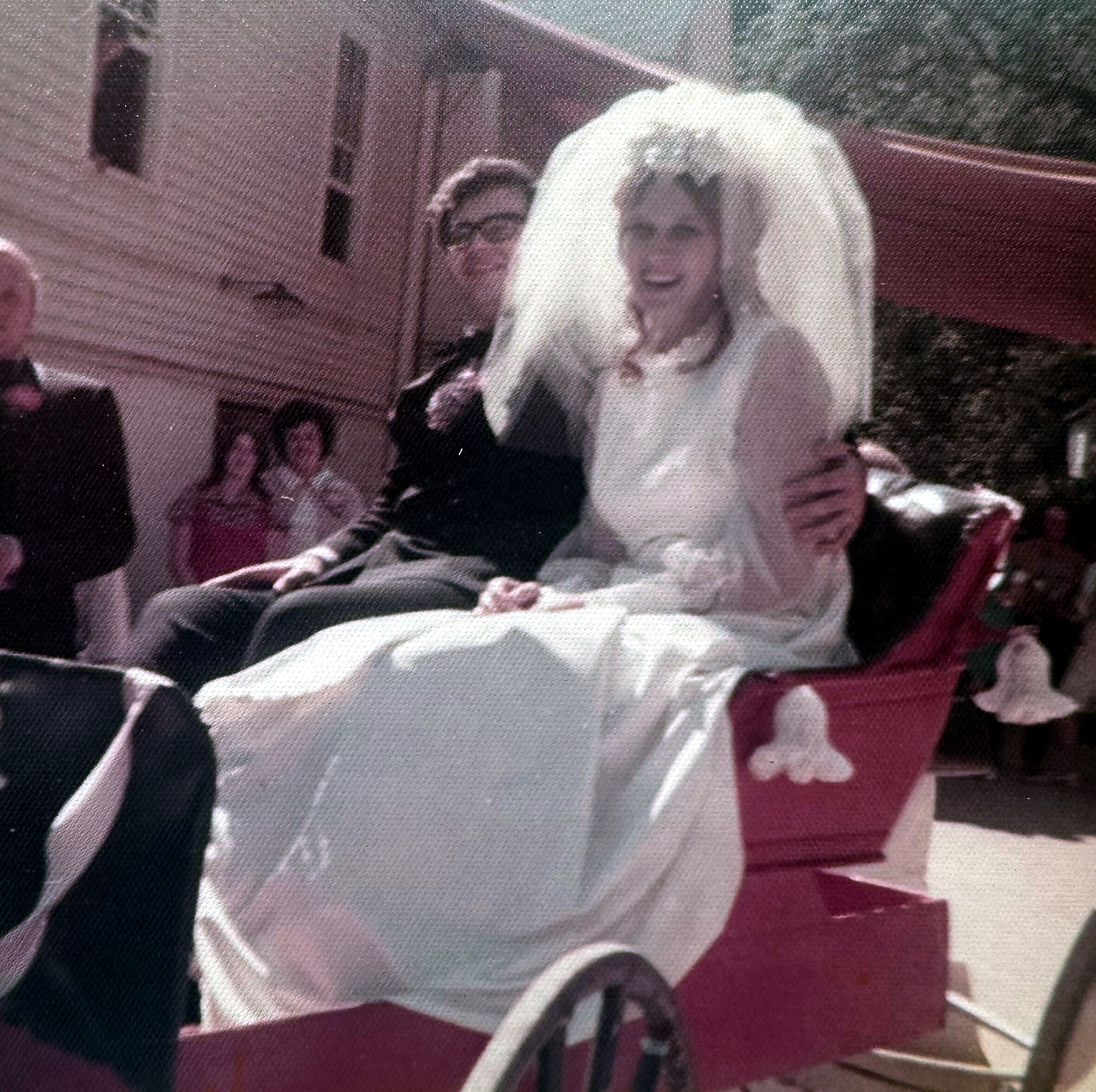 Bride in a white wedding dress and veil, smiling, seated next to a man with glasses indoors.
