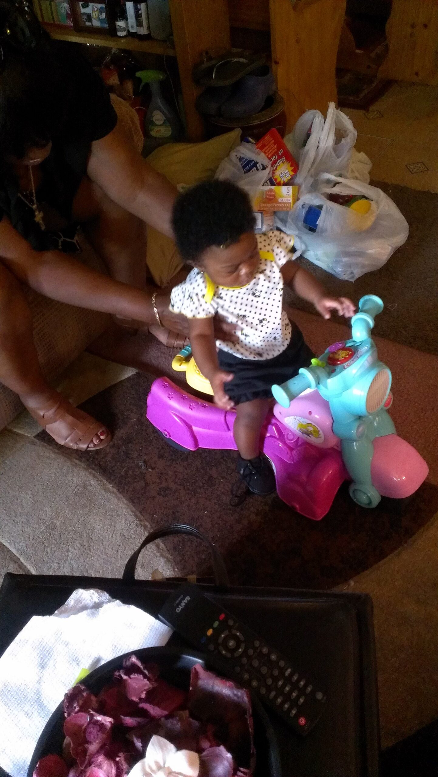 Young child sits on a colorful ride-on toy bike indoors, with an adult helping steady them.