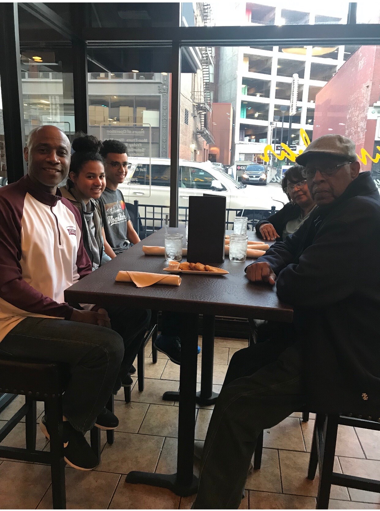 Six people sit at a cafe table by a large window; bread, water glasses, and a menu stand on the table, with an urban street outside.
